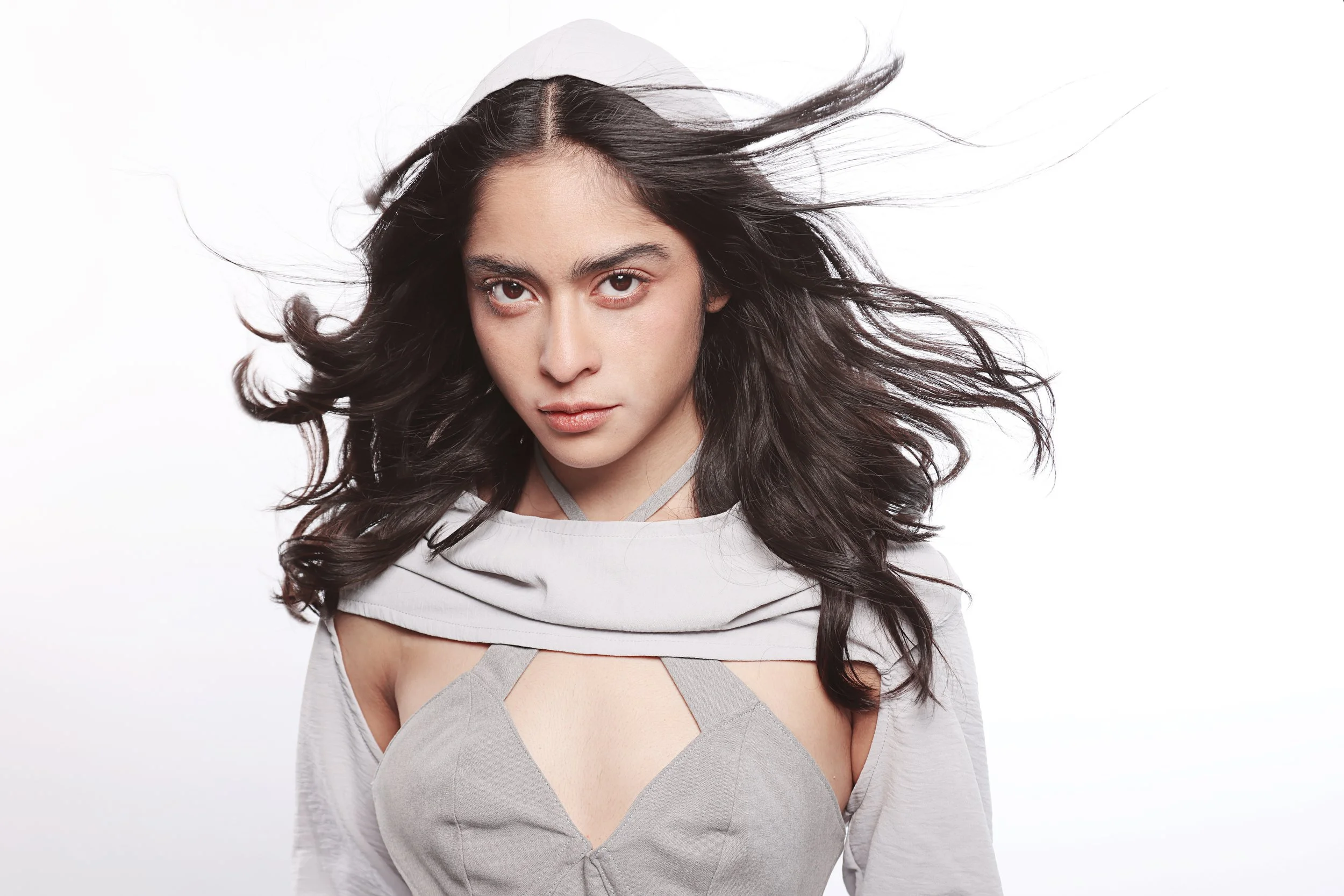 A woman with long dark hair, wearing a light gray top with cutouts, appears to be blowing gently with wind blowing her hair, against a plain white background.