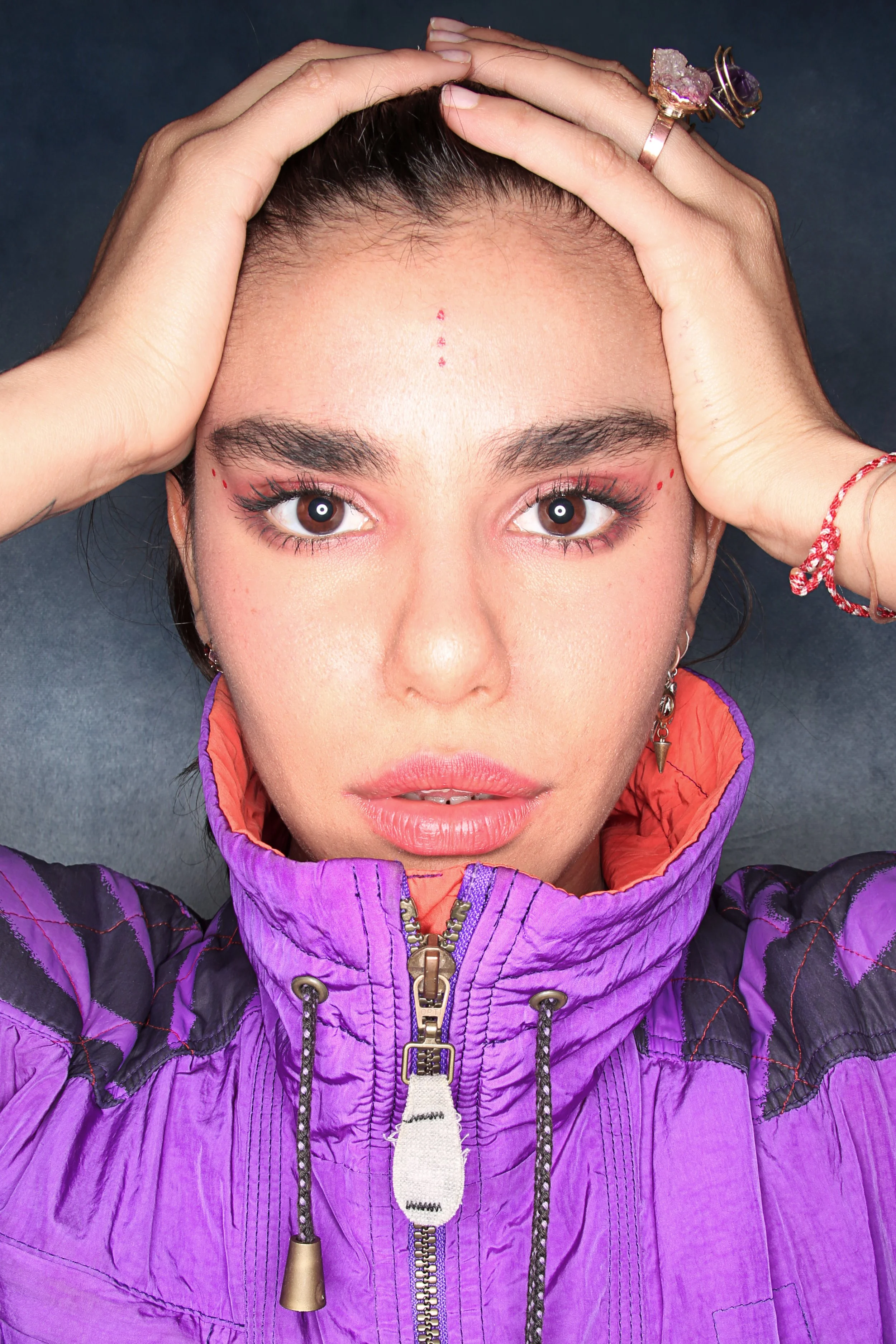A young woman with dark hair, brown eyes, and wearing makeup, lies on her back with hands on her head. She is wearing multiple rings and a red string bracelet on her left wrist. She is dressed in a purple puffy jacket with orange lining, and the phot