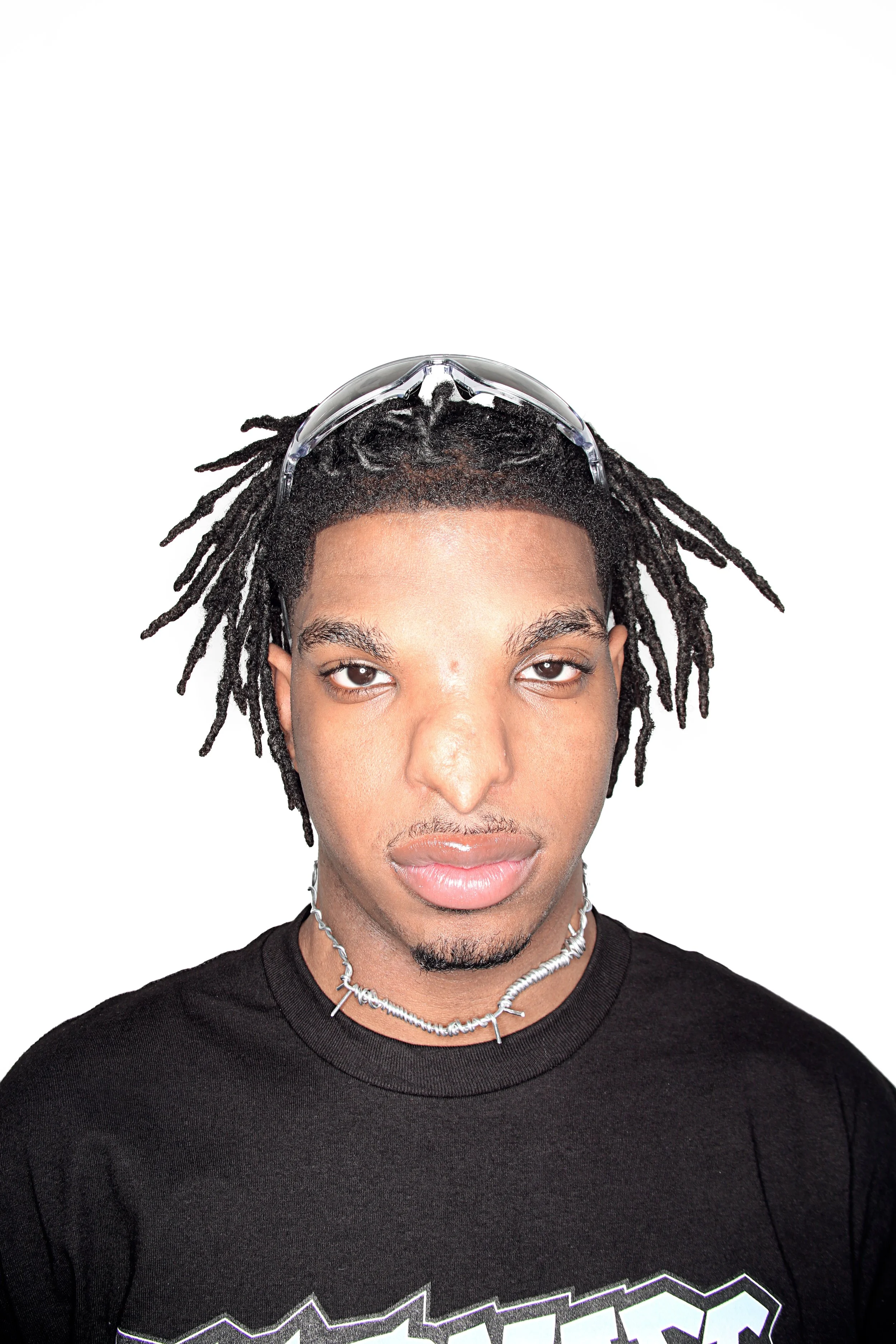 A young man with dreadlocks wearing safety goggles on his forehead, a black T-shirt with a graphic design, and layered silver necklaces, standing against a plain white background.