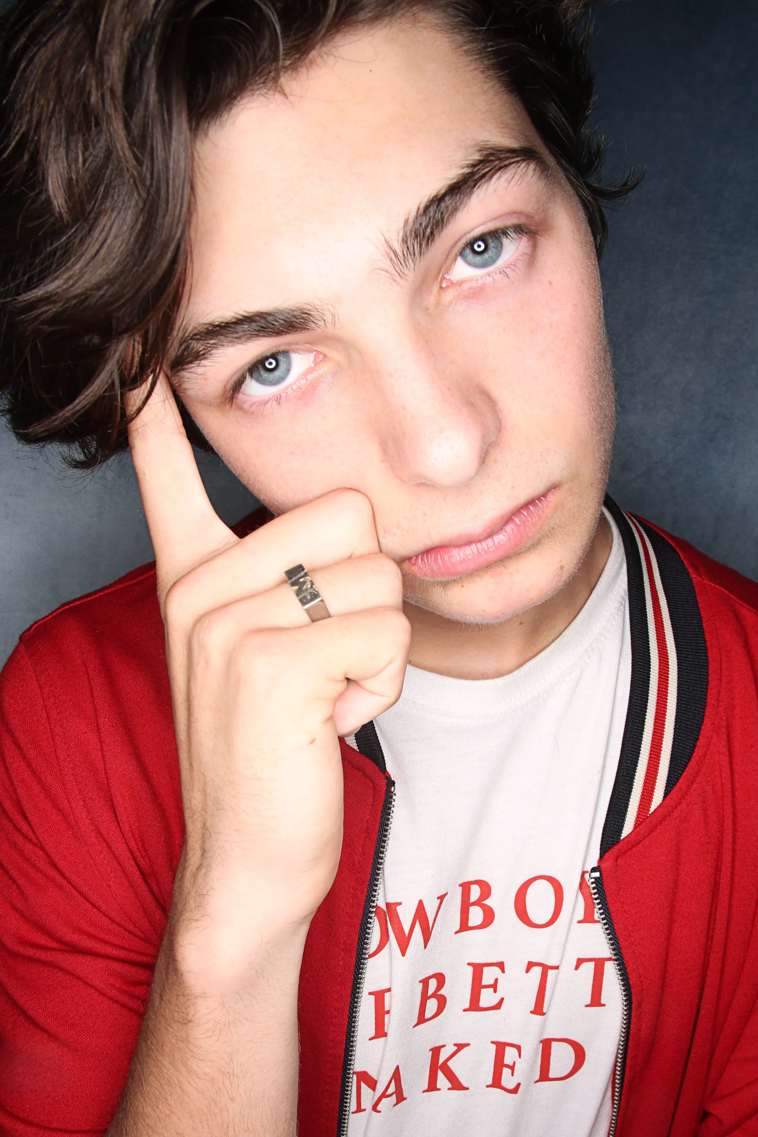 Close-up of a young man with blue eyes and dark hair, wearing a red jacket over a white t-shirt with red text. He is touching his face with his right hand, wearing a ring. The background is dark gray.