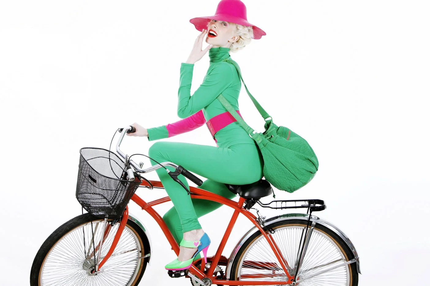 A woman wearing a bright pink hat, green bodysuit, and high heels, sitting sideways on a red bicycle with a black basket, against a white background.
