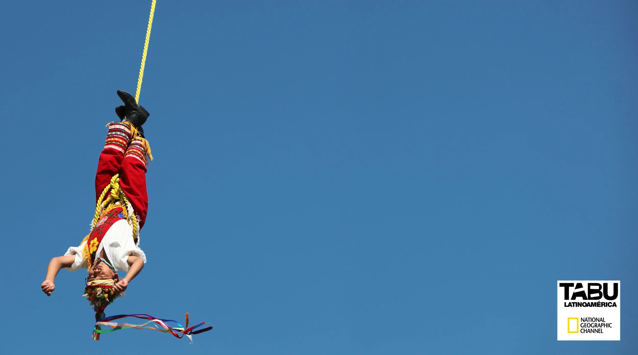 A person dressed in colorful, traditional Mexican attire performing aerial silk acrobatics against a clear blue sky. The performer is upside down with legs wrapped in vibrant fabric, holding onto yellow ropes. There are decorative ribbons at the bott
