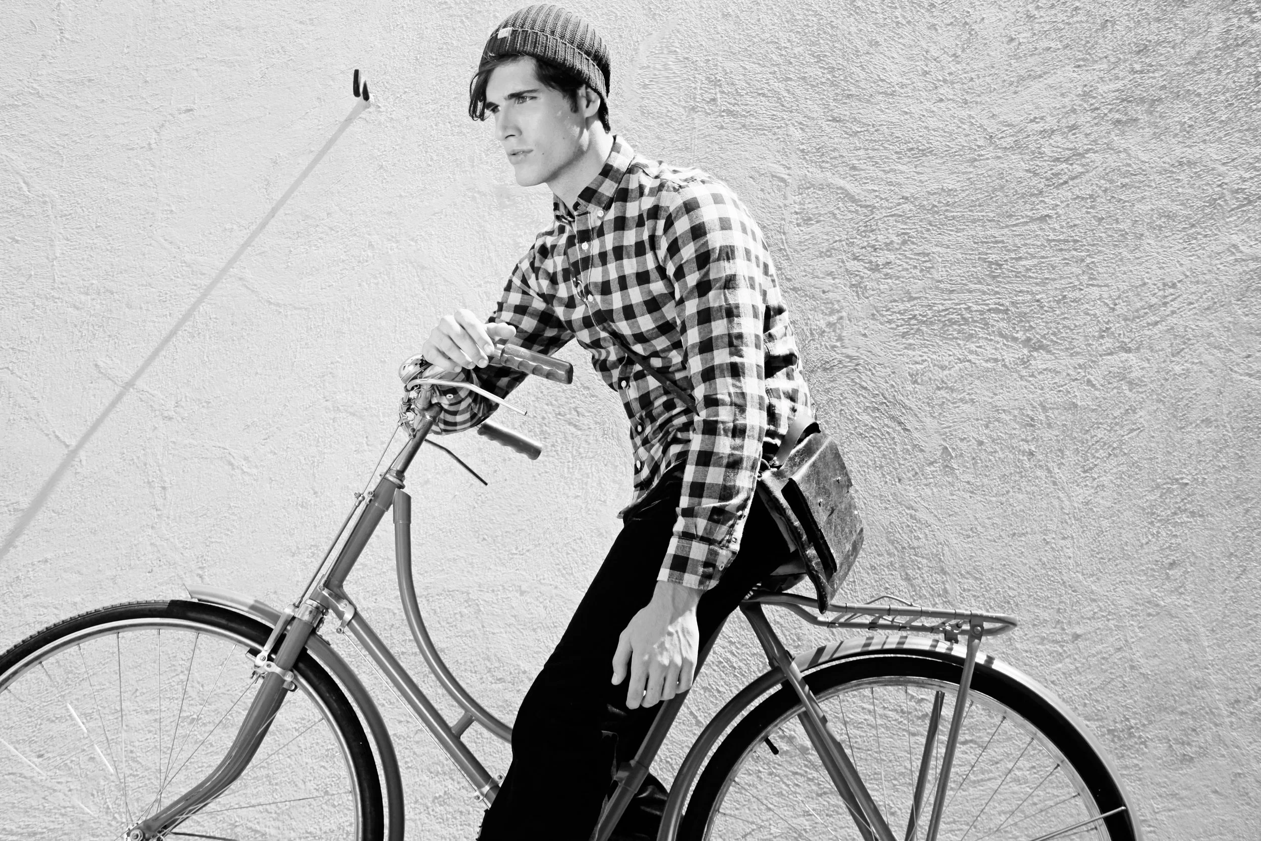 A young man wearing a checkered shirt, beanie, and dark pants, with a backpack, riding a bicycle against a textured wall, looking into the distance.