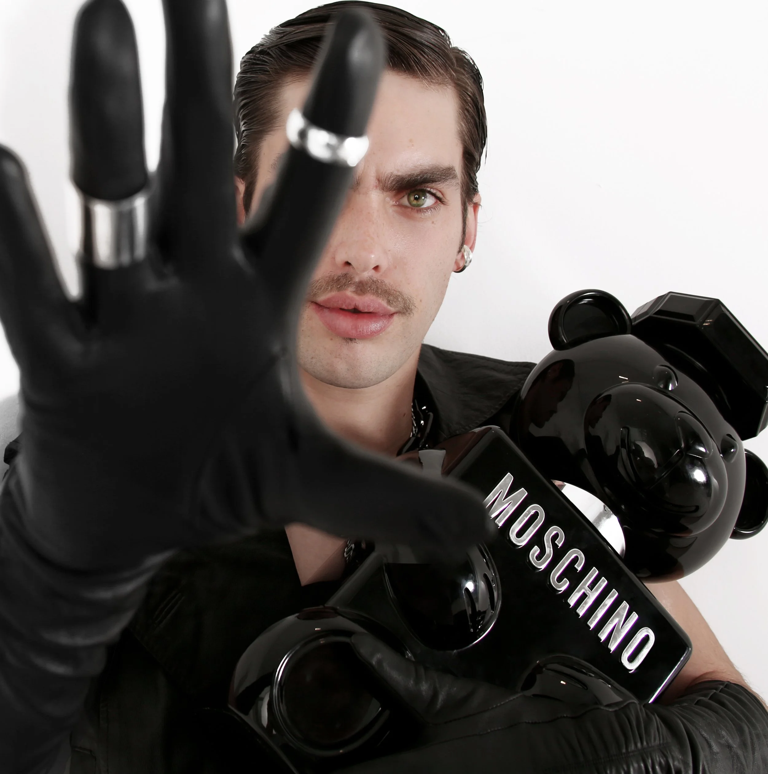 Close-up of a man with green eyes and slicked-back hair, partially obscured by black bottles with white or silver caps, and holding a black bottle with a purple label that reads 'MOSCHINO'.