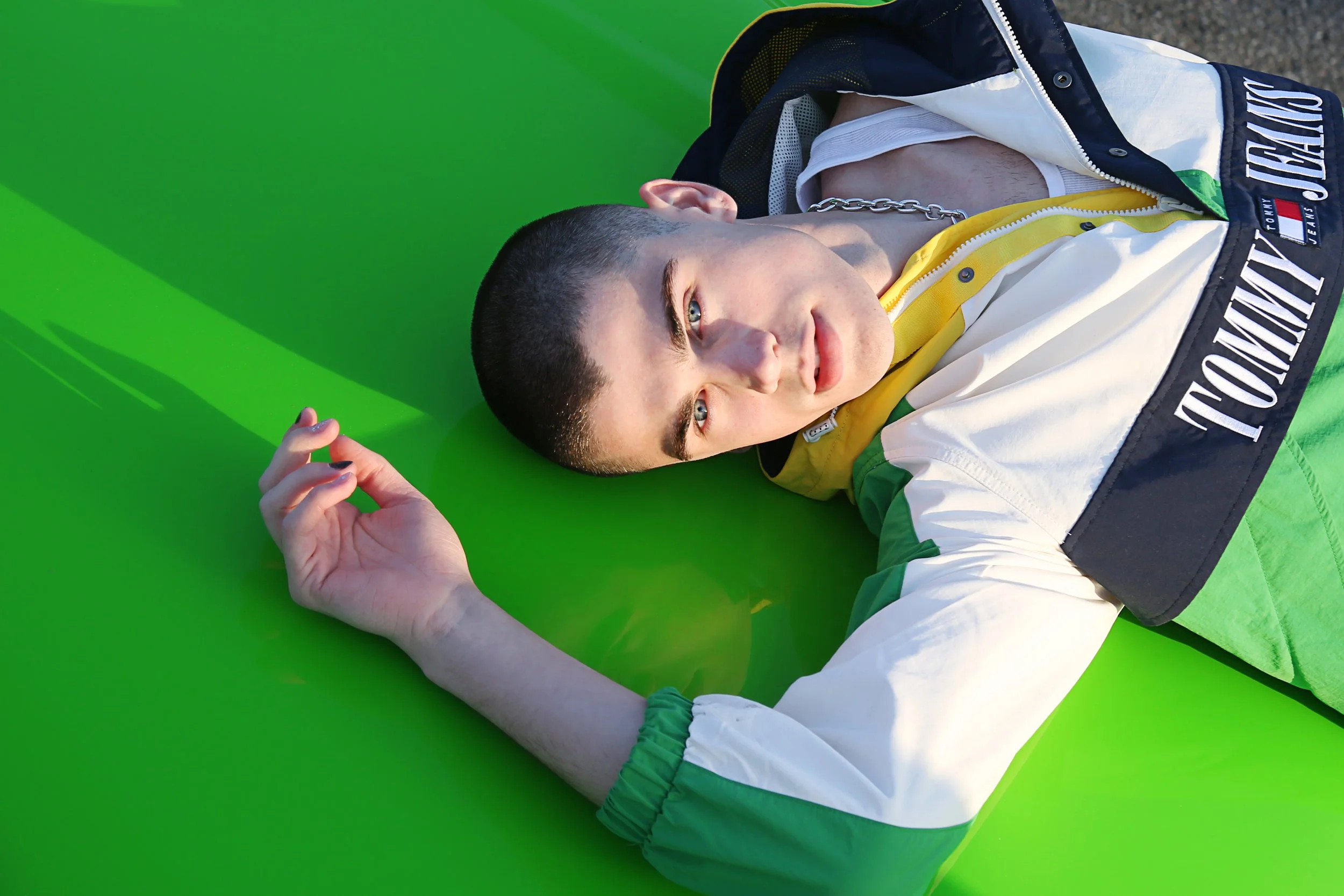 Young man with short dark hair, blue eyes, and light skin lying on a bright green mat outdoors, wearing a colorful jacket with yellow, white, and navy blue sections, and a gold chain necklace.