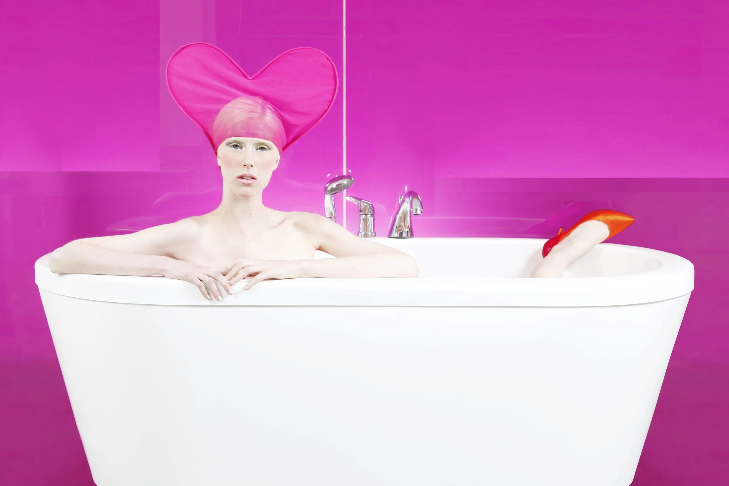 Person in a bathtub with pink head covering and heart-shaped pink hat, against a pink background.