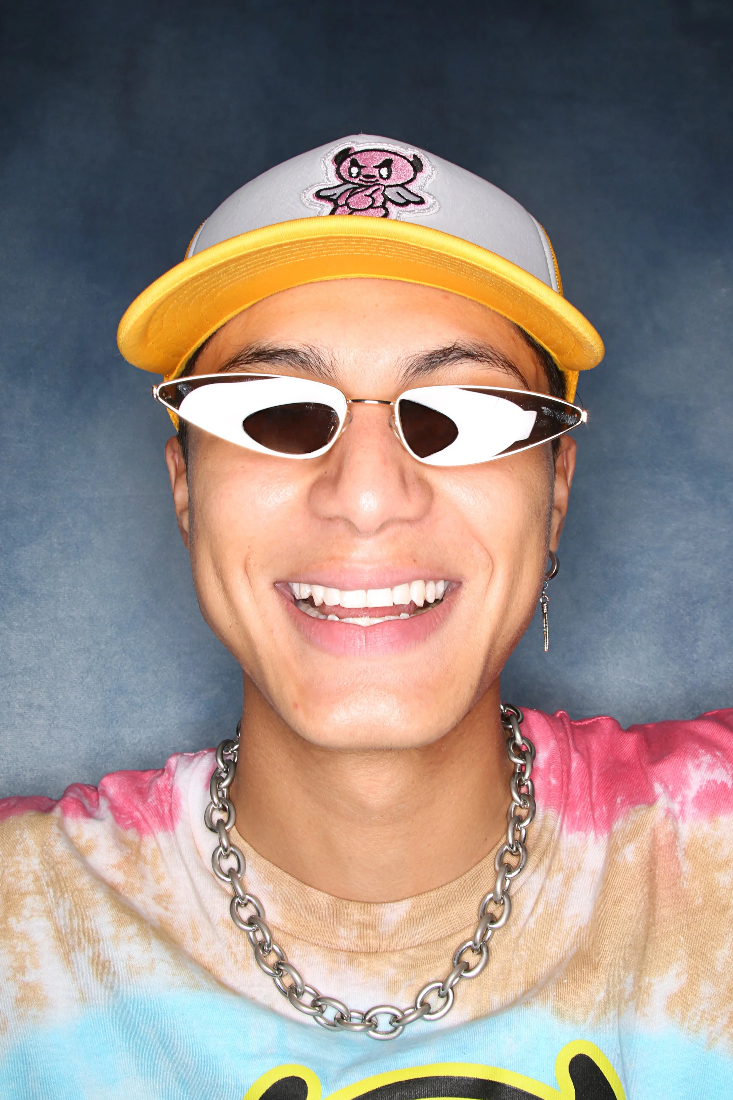 A person smiling with short hair wearing a yellow and gray cap with a pink cartoon animal embroidered on it, reflective sunglasses, a chunky silver chain necklace, and a tie-dye shirt with pink, orange, blue, and yellow colors under a blue background