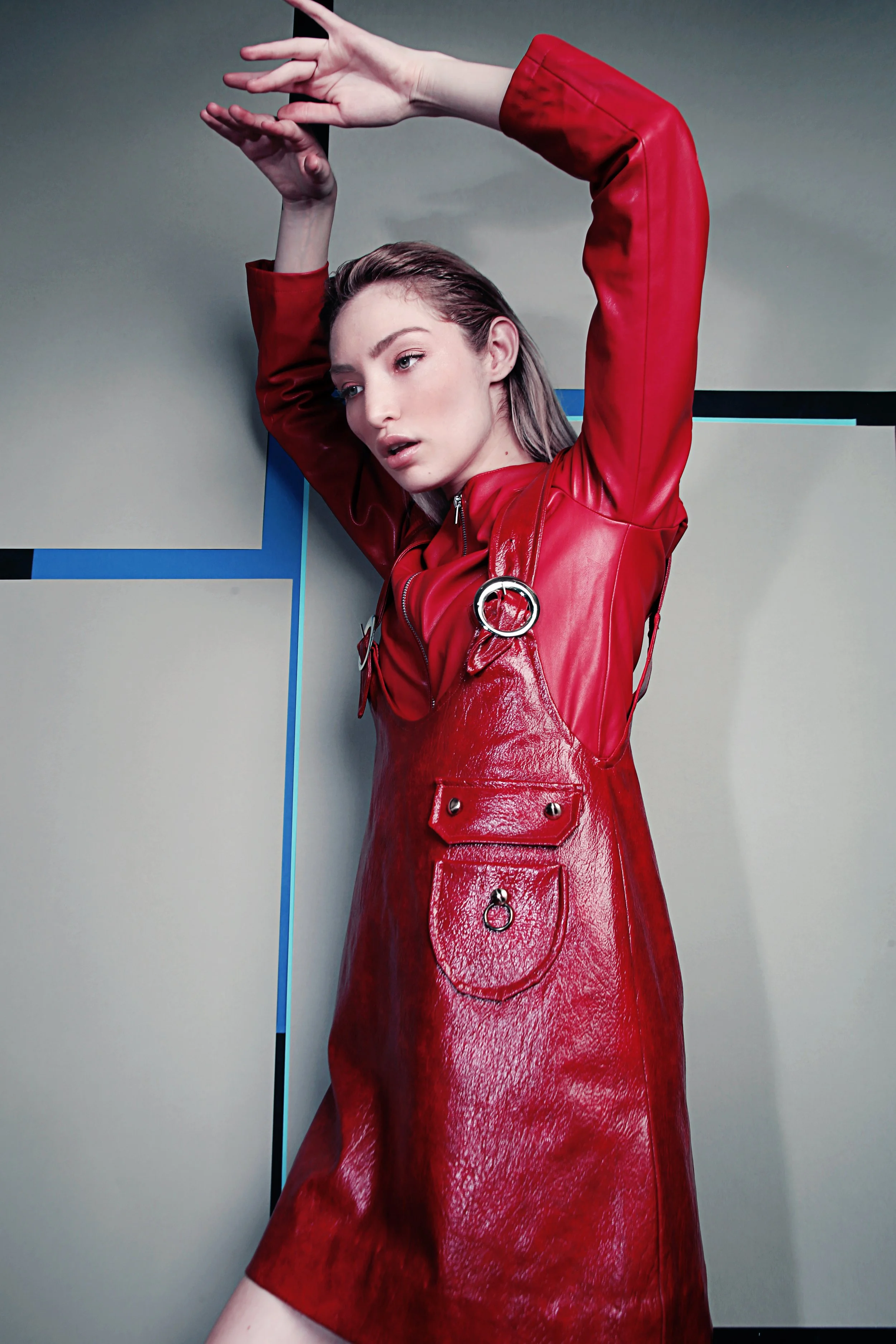 A woman with wavy hair wearing a shiny red leather dress with pocket details and ring accents, posing against a modern geometric background with her arms raised above her head.