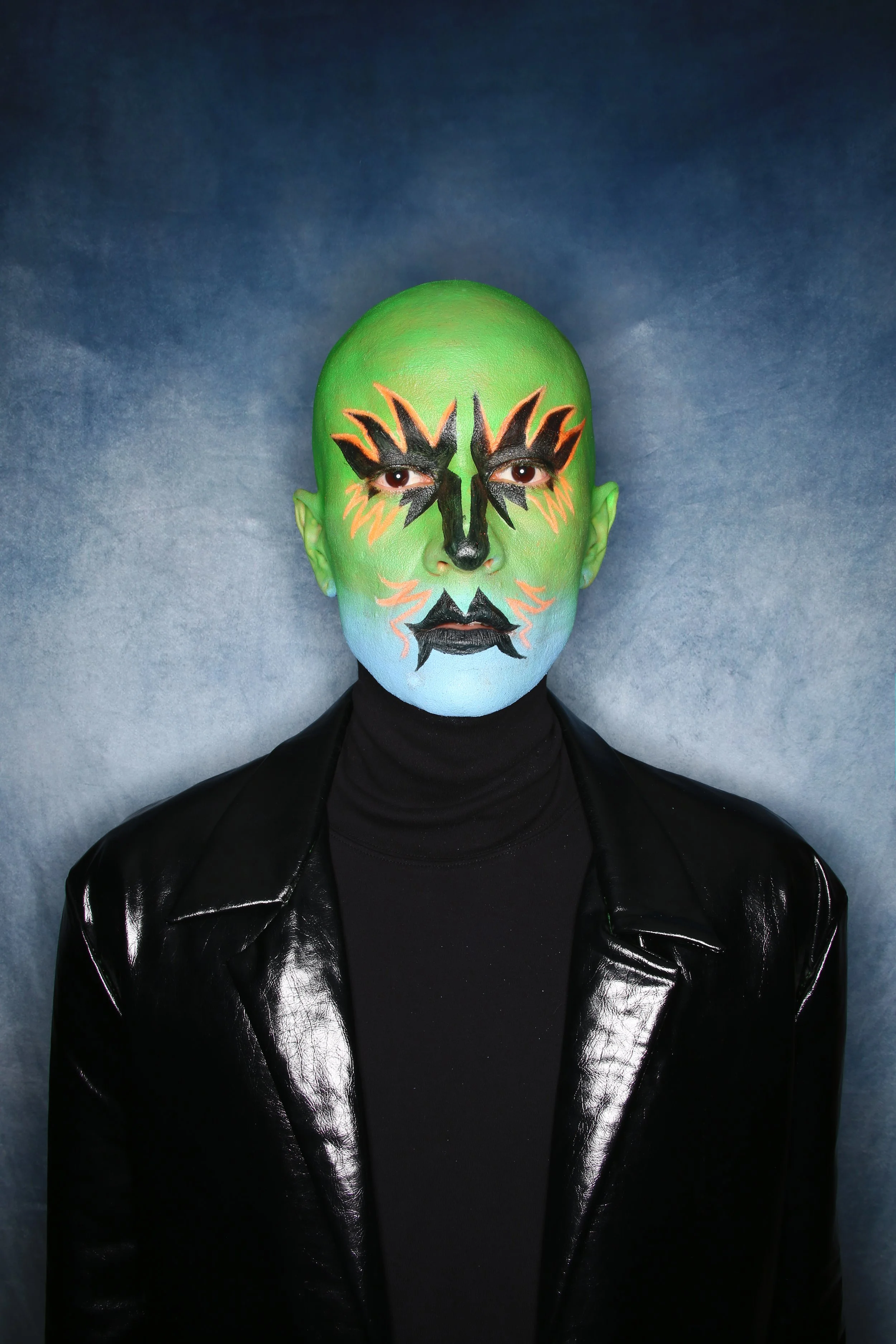 Person with bright green head, dramatic black and orange face makeup, wearing a black turtleneck and shiny black leather jacket, against a blue-gray background.