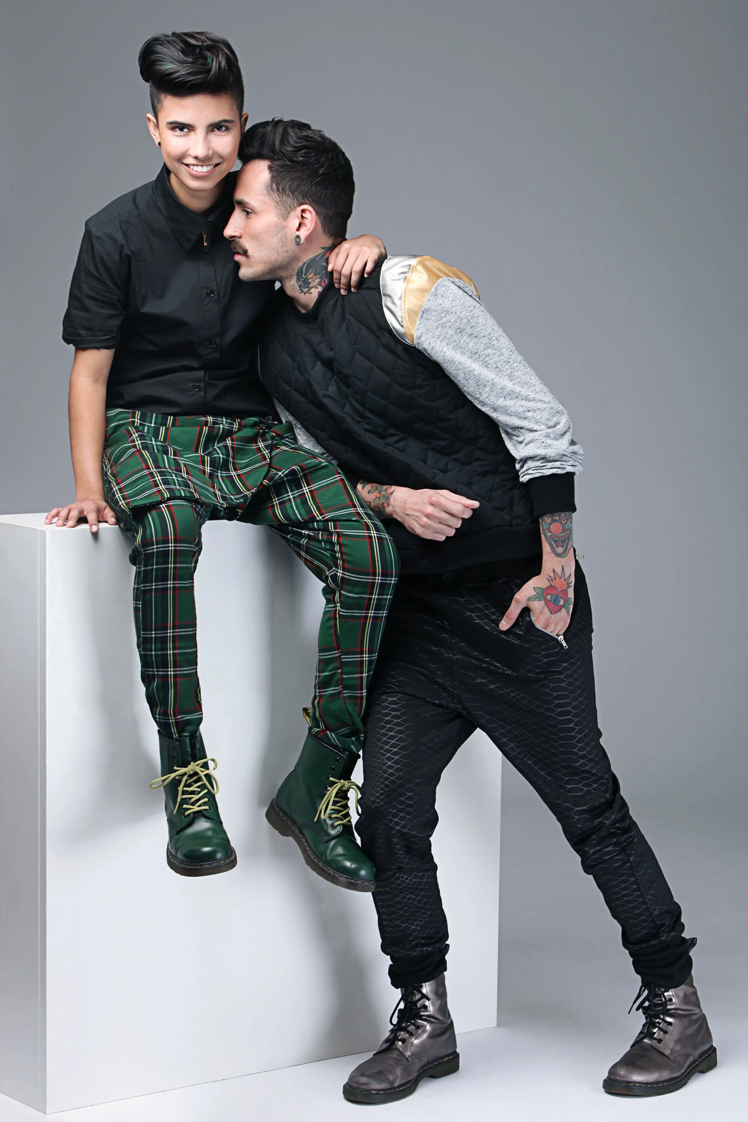 Two individuals with tattoos and dark hair posing against a plain gray backdrop. One is seated on a white block, wearing a black shirt, plaid pants, and green boots; the other is standing, leaning to the seated person, wearing a gray sweatshirt, blac