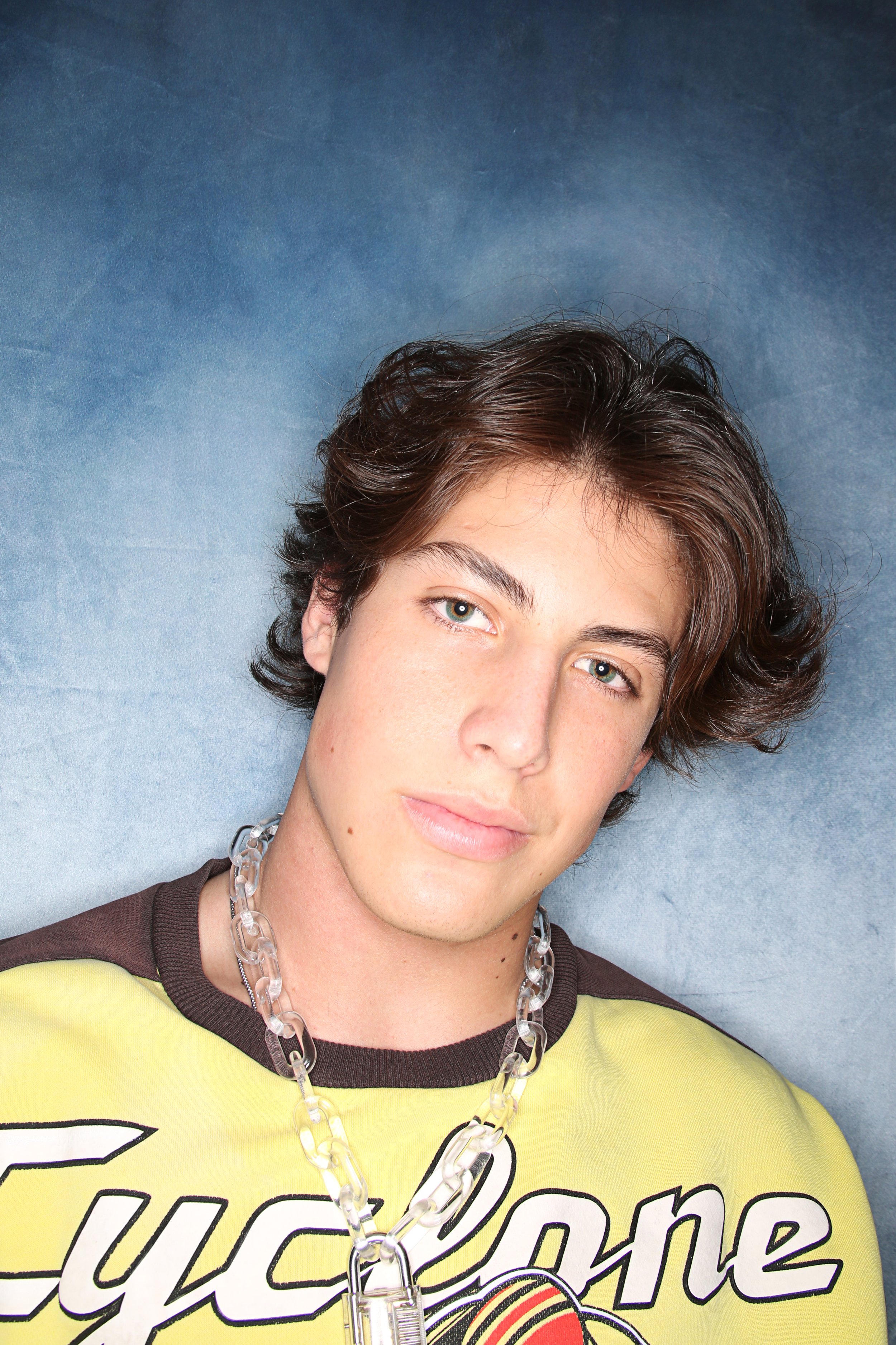 A person with short wavy brown hair and light skin, wearing a yellow shirt with black trim, a chunky clear plastic chain necklace, and a black and white graphic design, standing against a blue background.