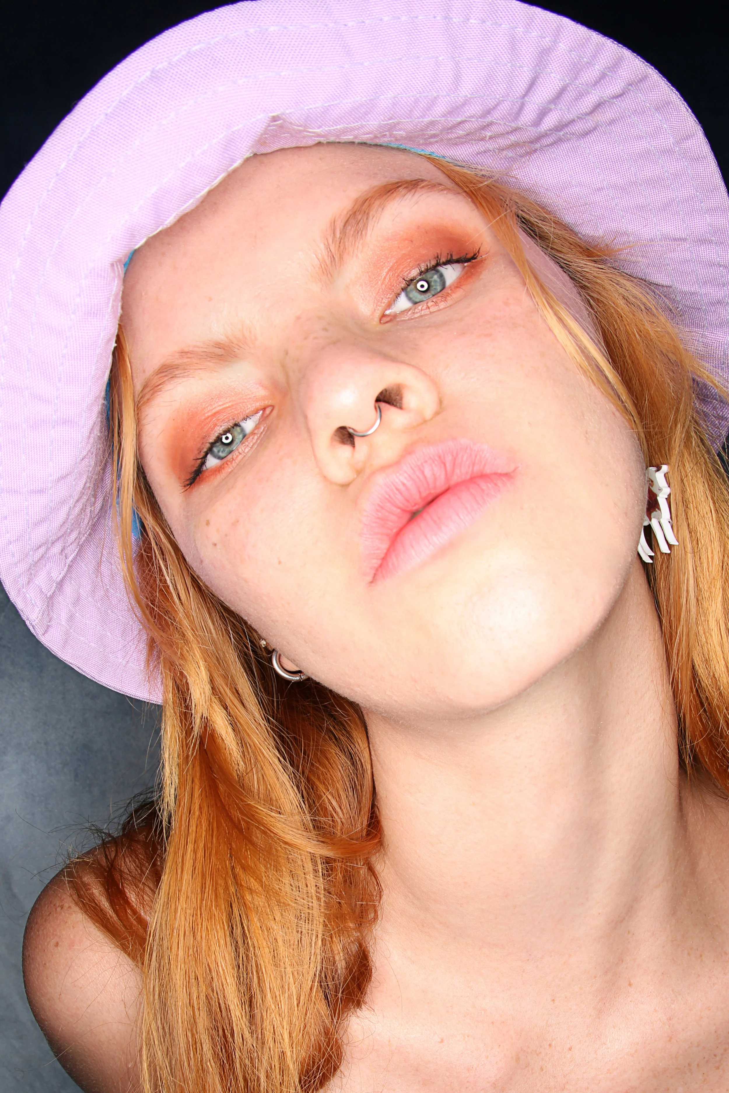 Close-up of a woman with red hair, wearing a lavender sunhat, looking at the camera with relaxed expression, against a dark background.