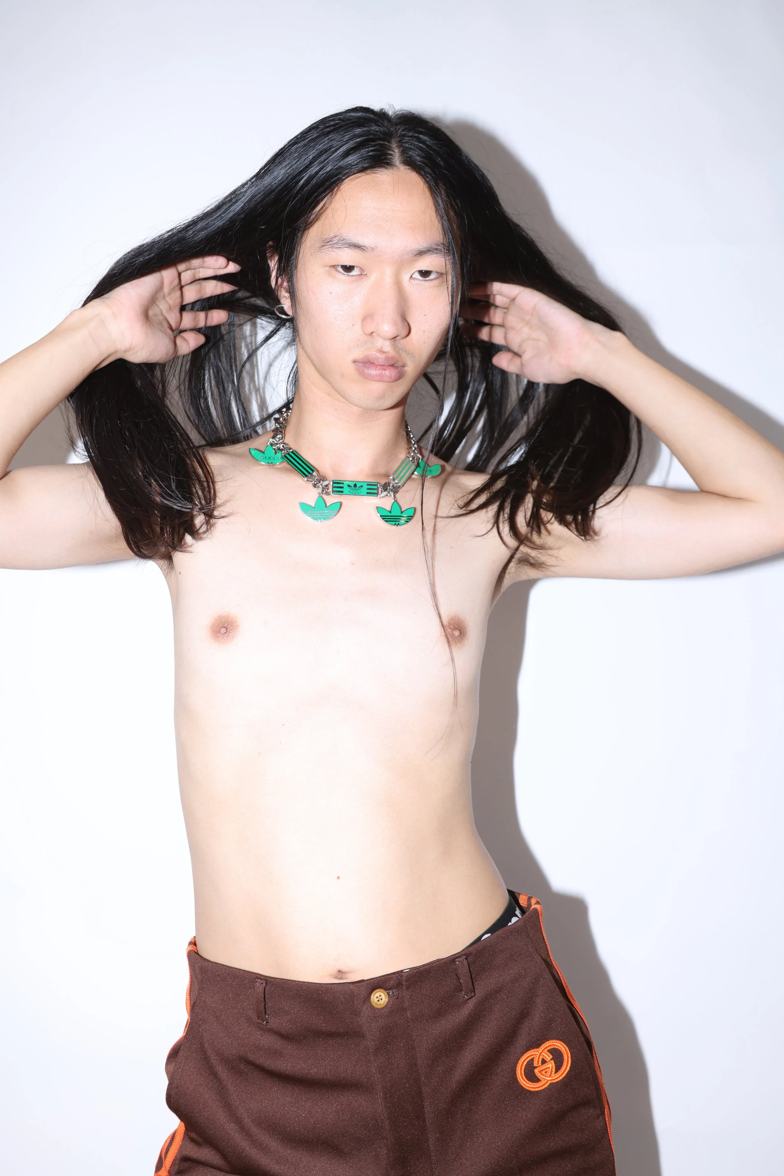 A shirtless young man with long black hair holds his hair back with both hands, wearing a chunky chain necklace with green, blue, and black accents, and brown pants with orange piping and an orange Gucci logo.