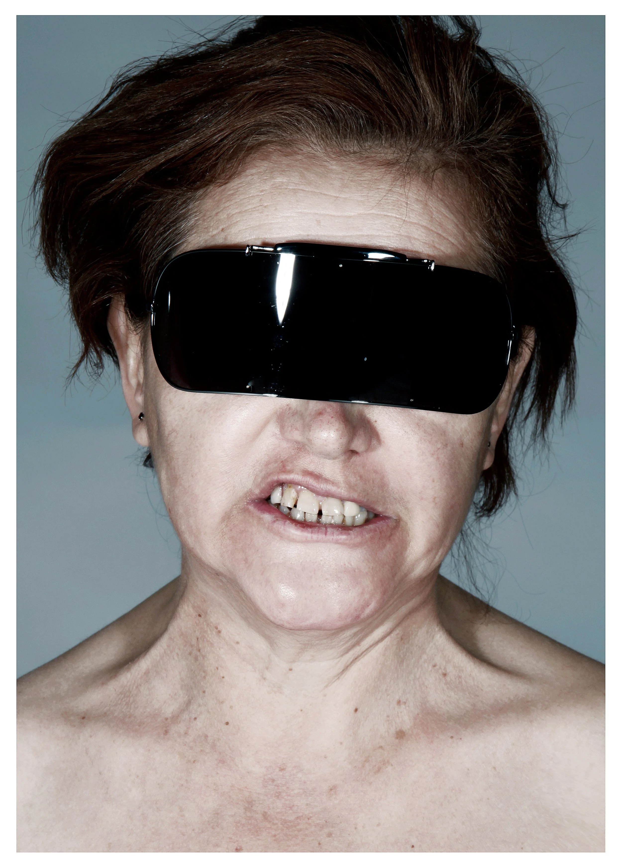A close-up of a woman with brown hair, wearing large black sunglasses, smiling, with visible facial wrinkles and a small earring on her left ear. Her neck and shoulders are bare, showing skin with some freckles and age spots.