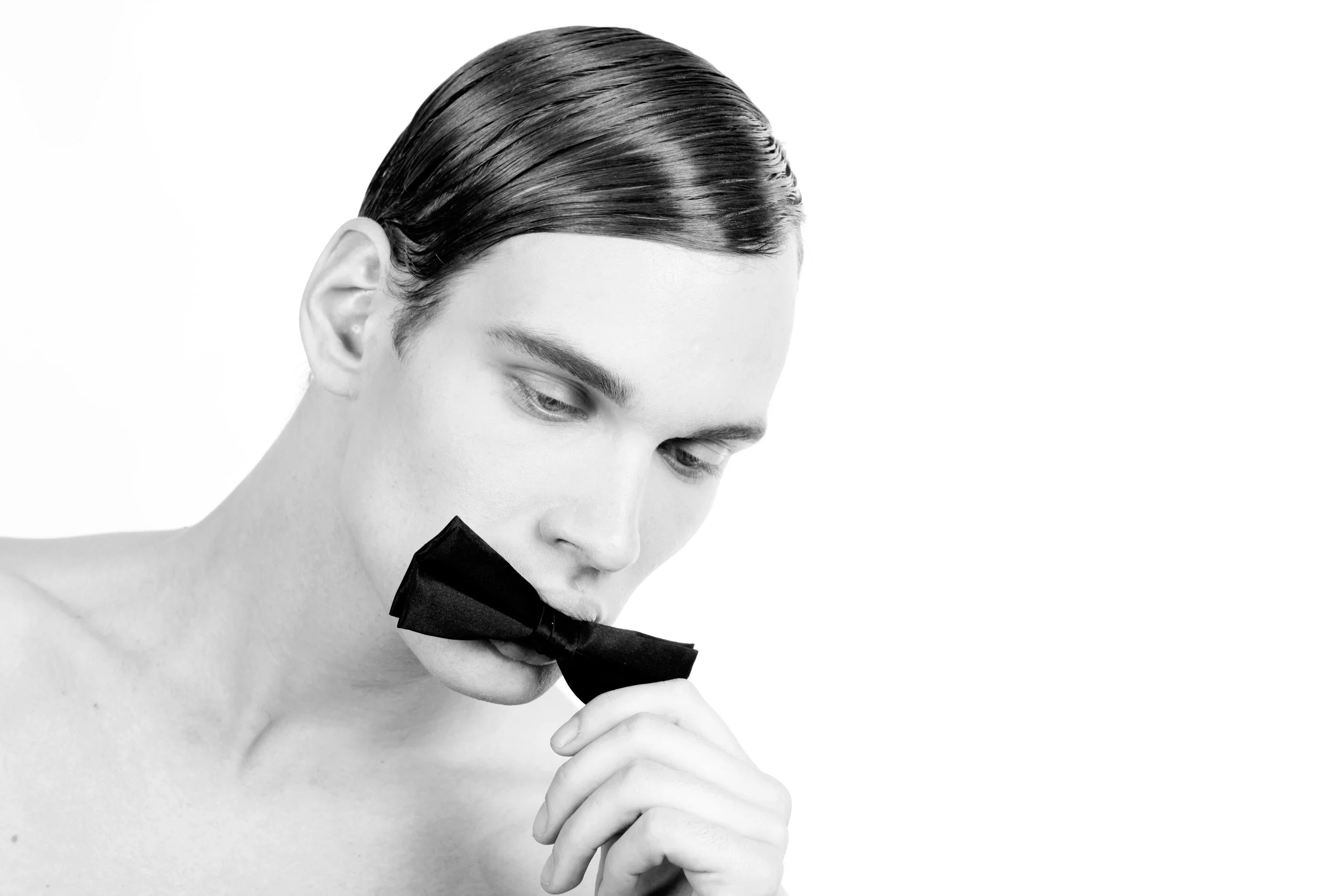 A black and white image of a young man with neatly combed hair, holding a black bow tie near his lips, with a contemplative expression.