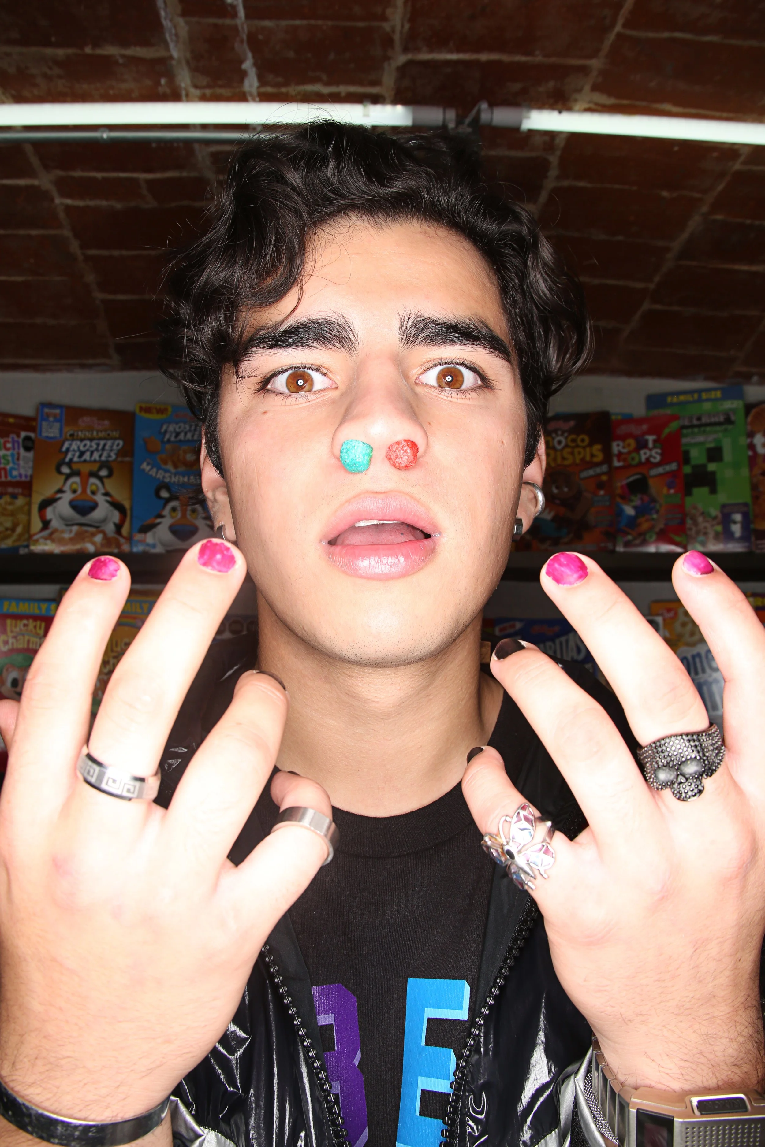 A young person with dark, curly hair and brown eyes, holding up their hands with painted nails and wearing rings and a skull ring. They are wearing a black shirt and a shiny black jacket with a zipper. There are cereal boxes on shelves behind them, i