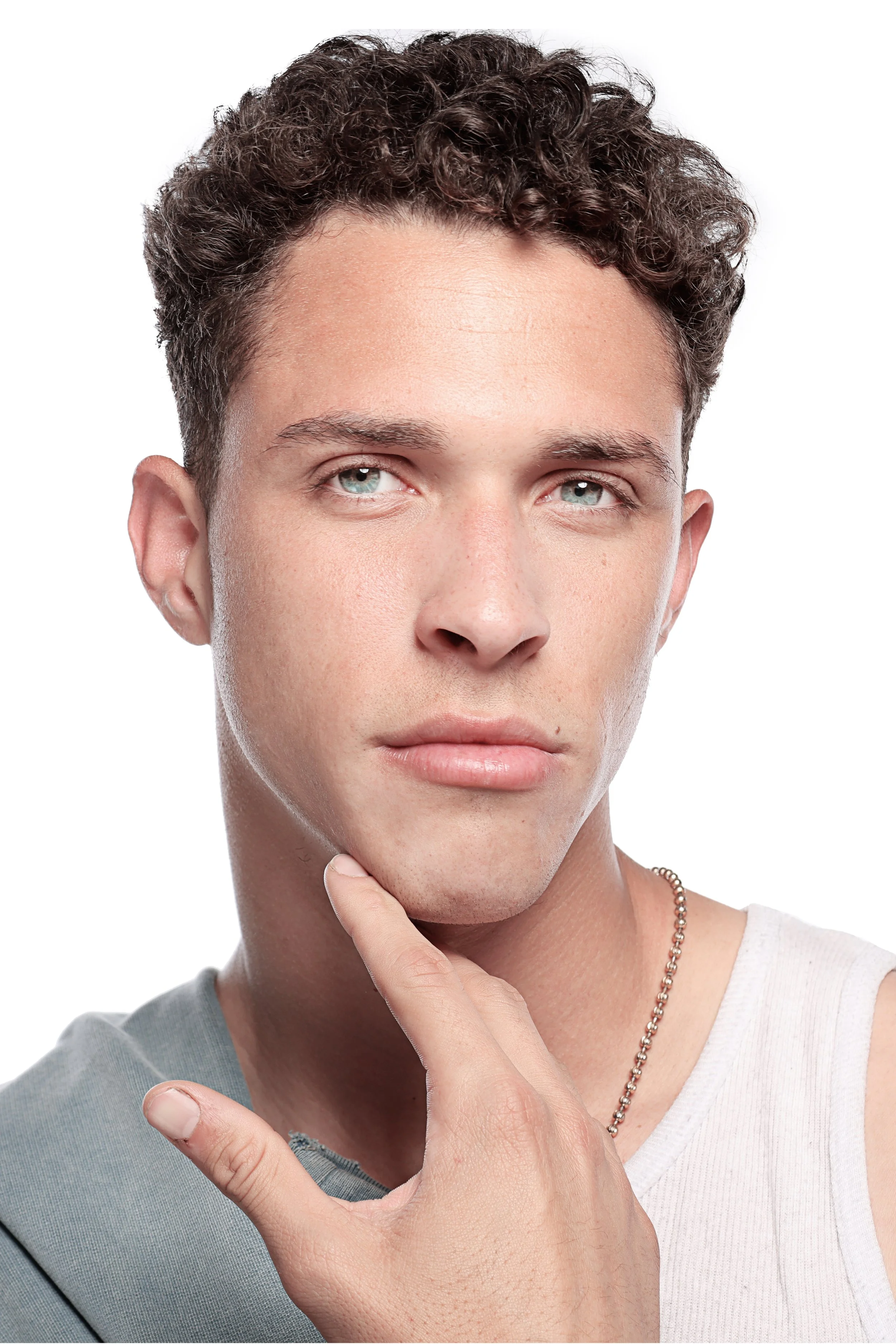 Close-up of a woman with short, curly dark hair, light blue eyes, holding her chin and index finger close to her lips, wearing a white top, and a silver chain necklace, with a neutral expression.