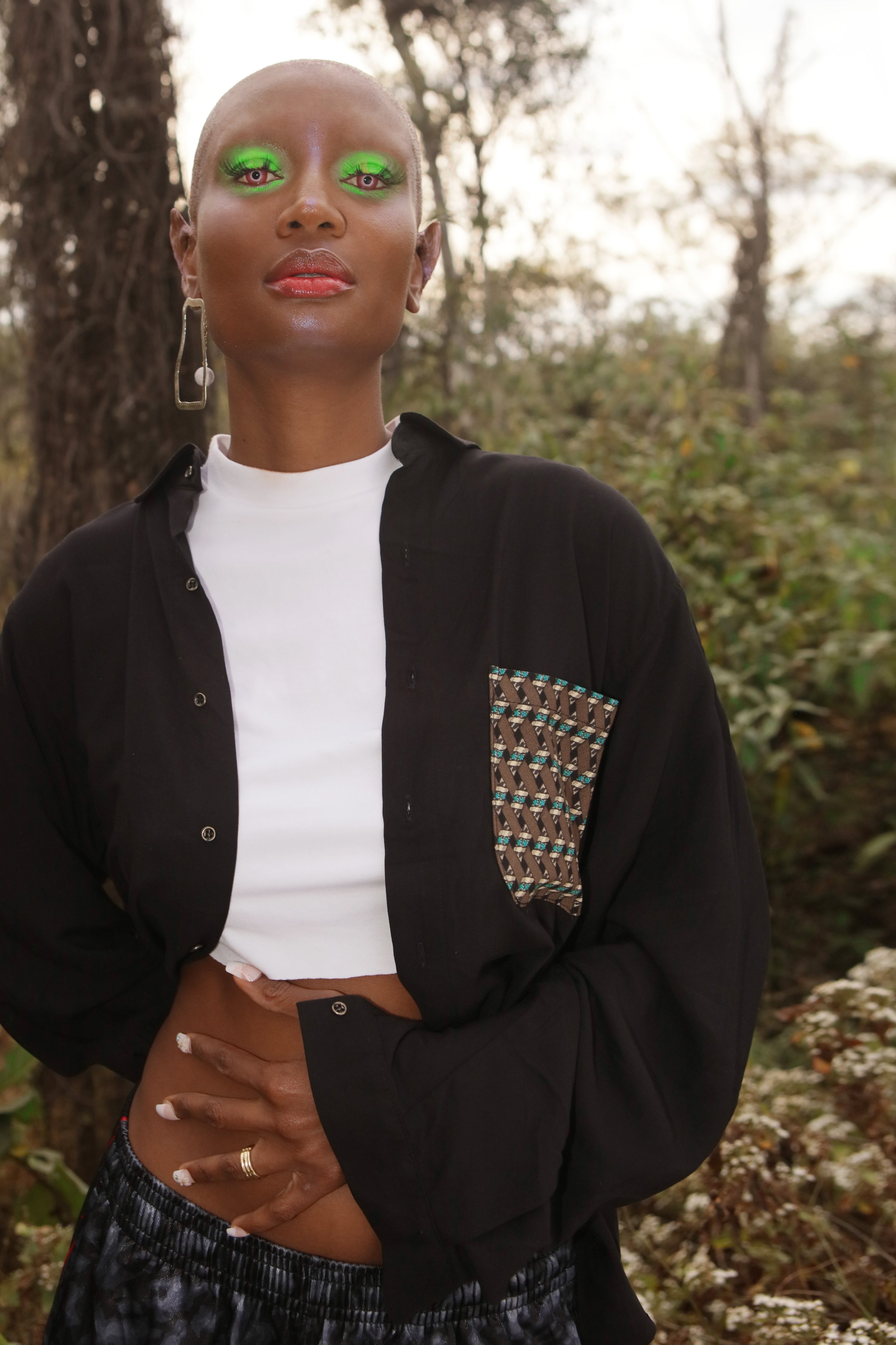 Close-up of a person with short platinum blonde hair, wearing bright green makeup on the eyelids, red lipstick, earrings, and a black jacket with a patterned pocket, standing outdoors with trees and foliage in the background.