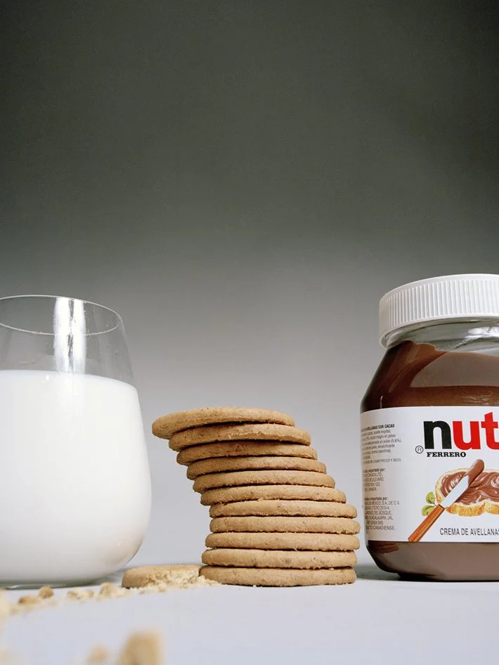 A glass of milk, a stack of cookies, and a jar of Nutella hazelnut spread on a white surface against a grey background.