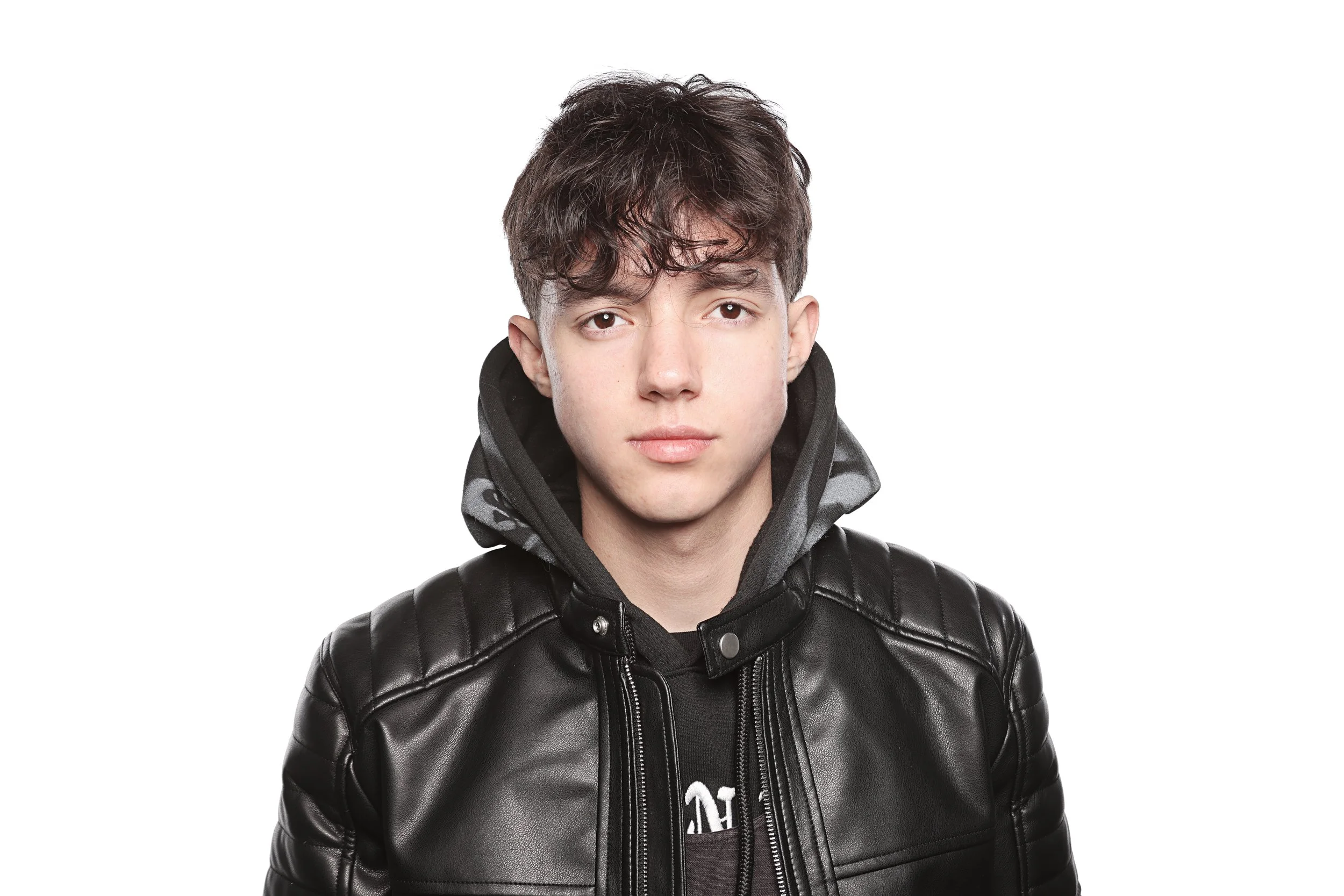 Young man with dark curly hair wearing a black leather jacket and hoodie, standing against a plain white background.