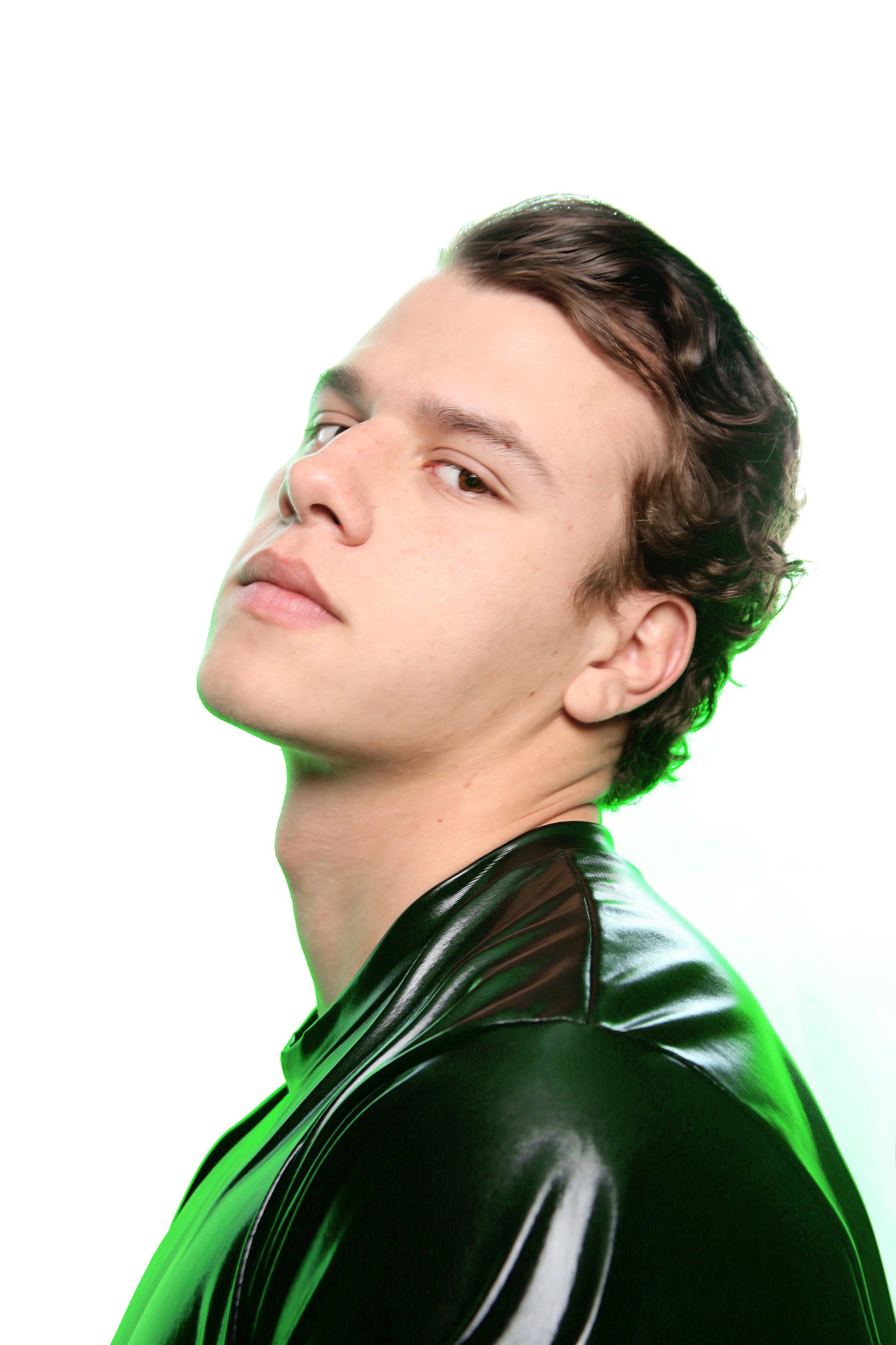 A young man with styled brown hair and light skin, looking slightly upward, wearing a shiny black leather jacket against a white background.
