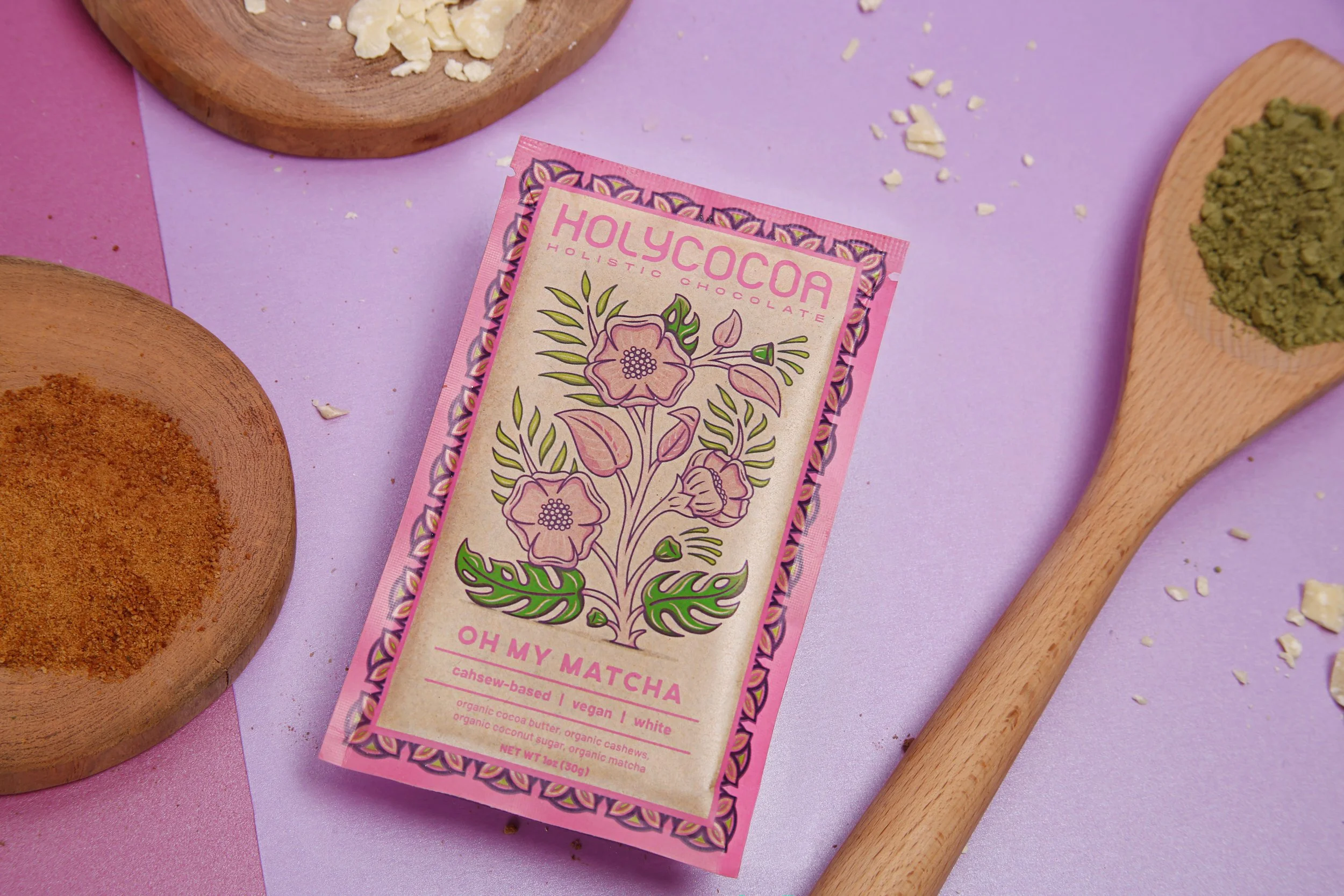A pink and beige box of Holy Cocoa holistic chocolate, featuring a floral illustration and text promoting its ingredients like organic cocoa butter and matcha, placed on a pastel pink and purple background with wooden bowls and a spoon containing gre