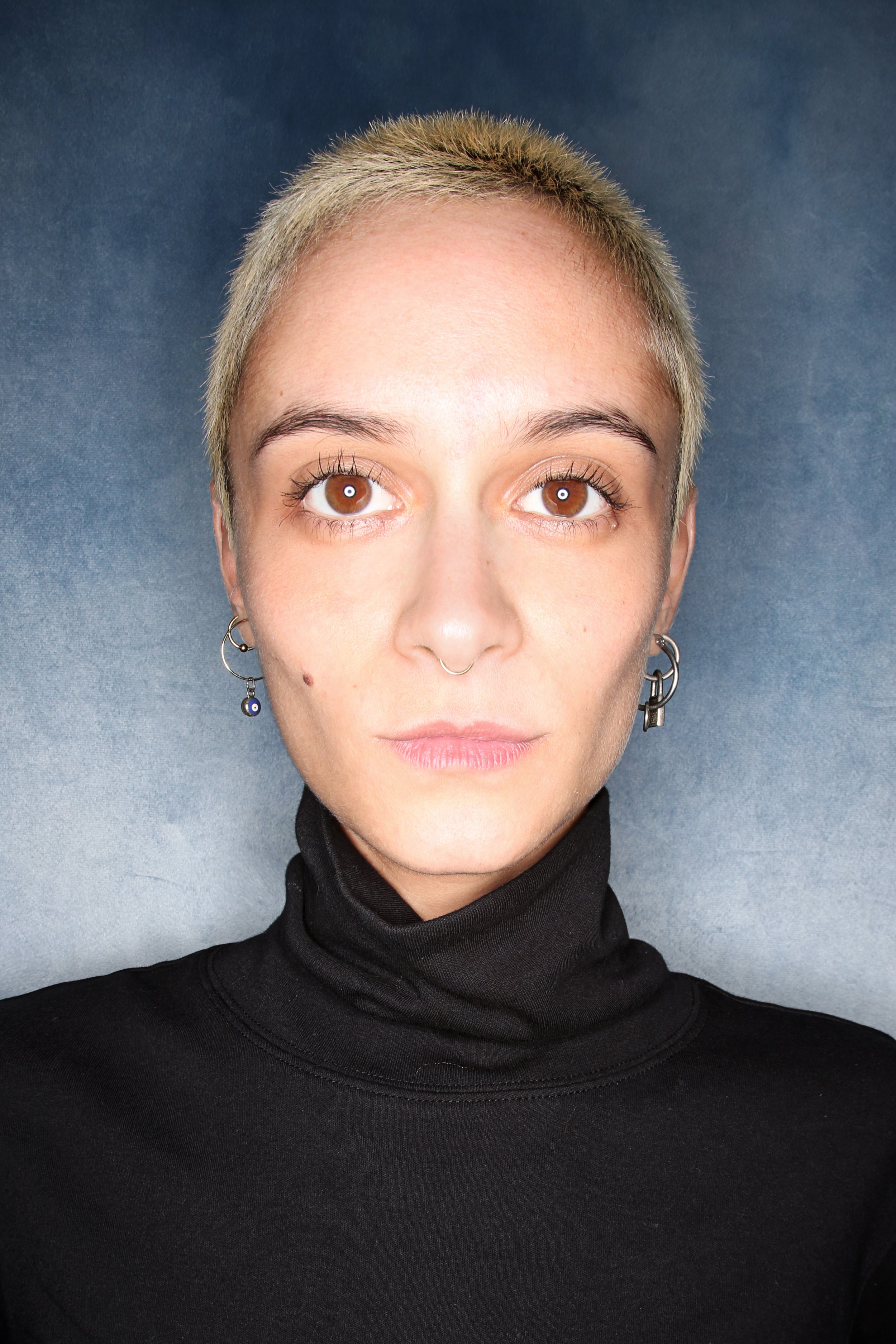 Close-up portrait of a young woman with short, platinum blonde hair, brown eyes, multiple earrings, septum piercing, and wearing a black turtleneck against a blue background.