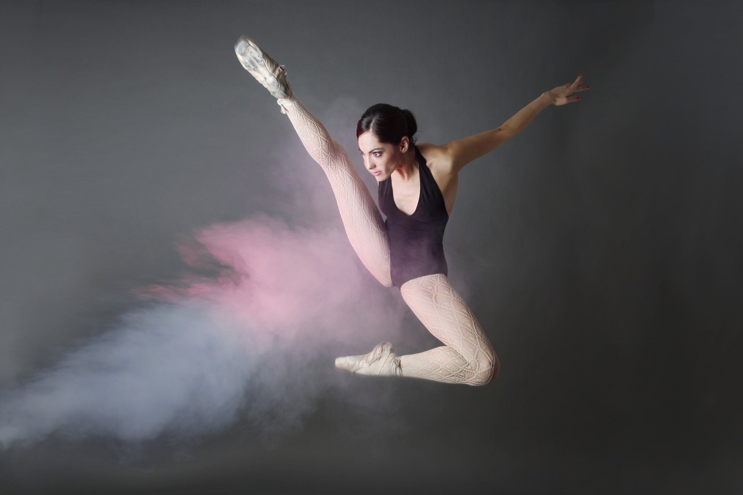 A female ballet dancer performing a high kick or leap against a gray background with pink and blue smoke effects.