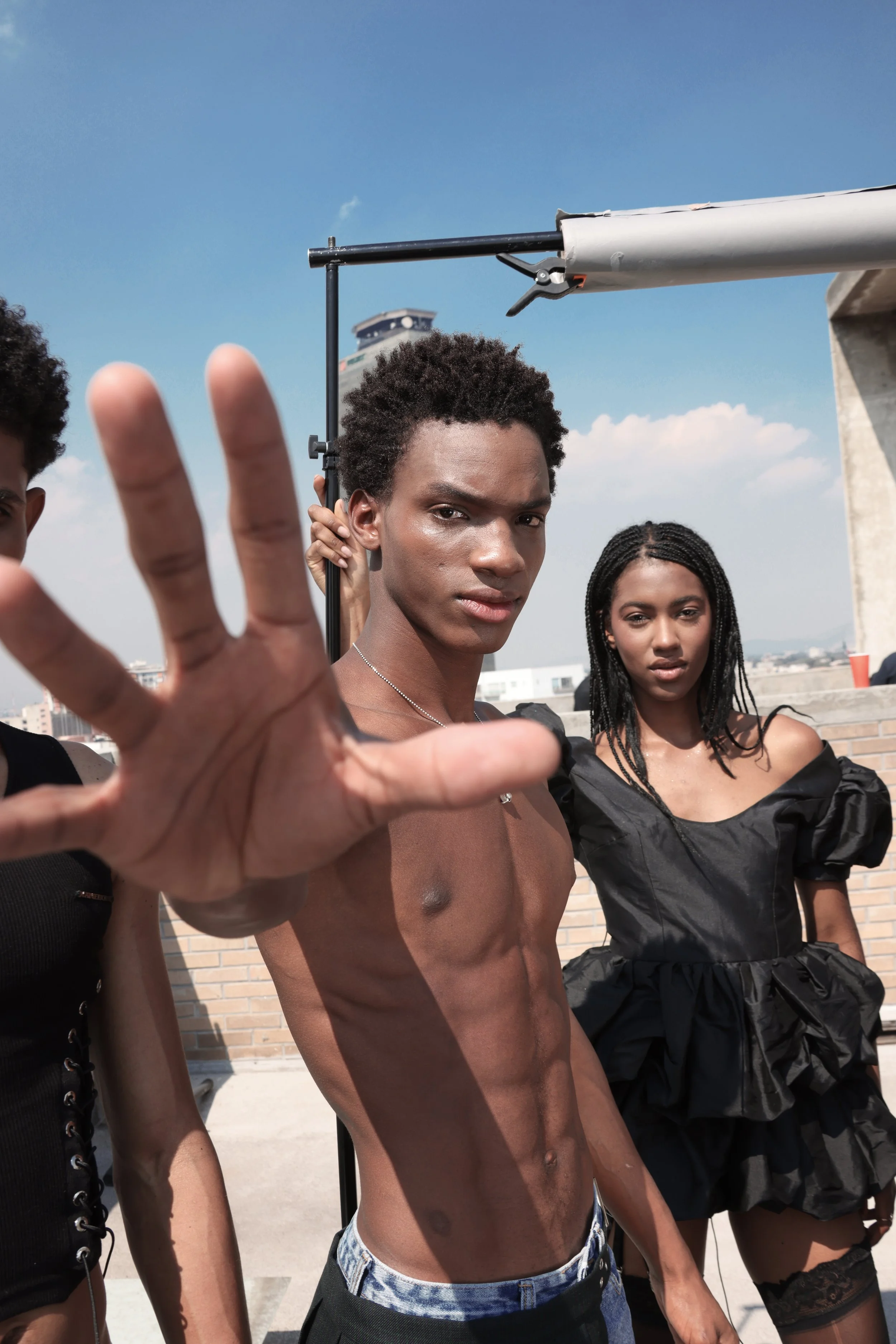 A shirtless young man with curly hair and a serious facial expression standing outside, with two women in black outfits, and a person holding a boom microphone in the background. The sky is blue with some clouds.