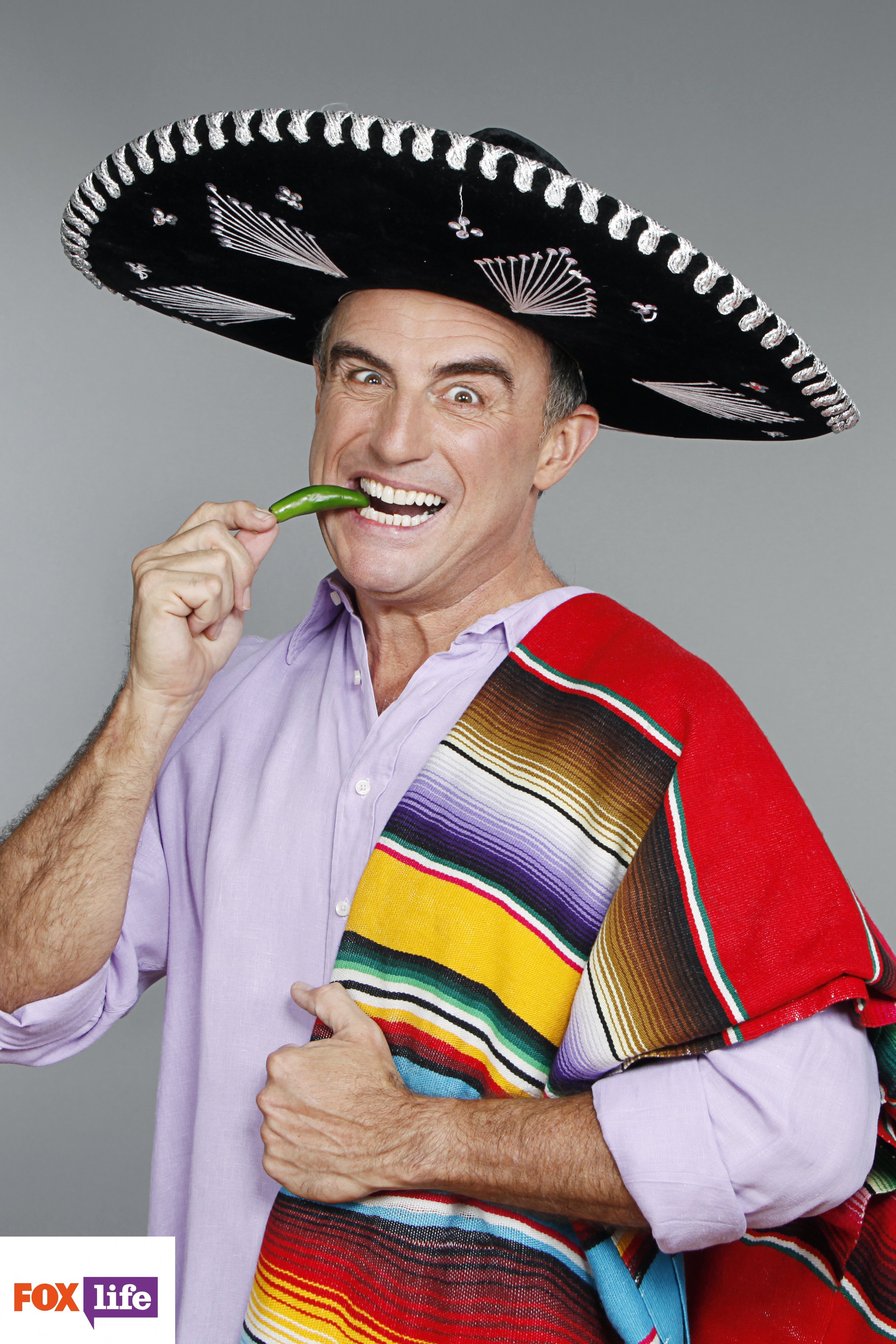 A man wearing a large traditional Mexican sombrero hat, holding a green chili pepper near his mouth, and smiling with wide eyes. He is dressed in a light purple shirt and a colorful striped shawl. The background is plain gray, and a logo of FOX Life 