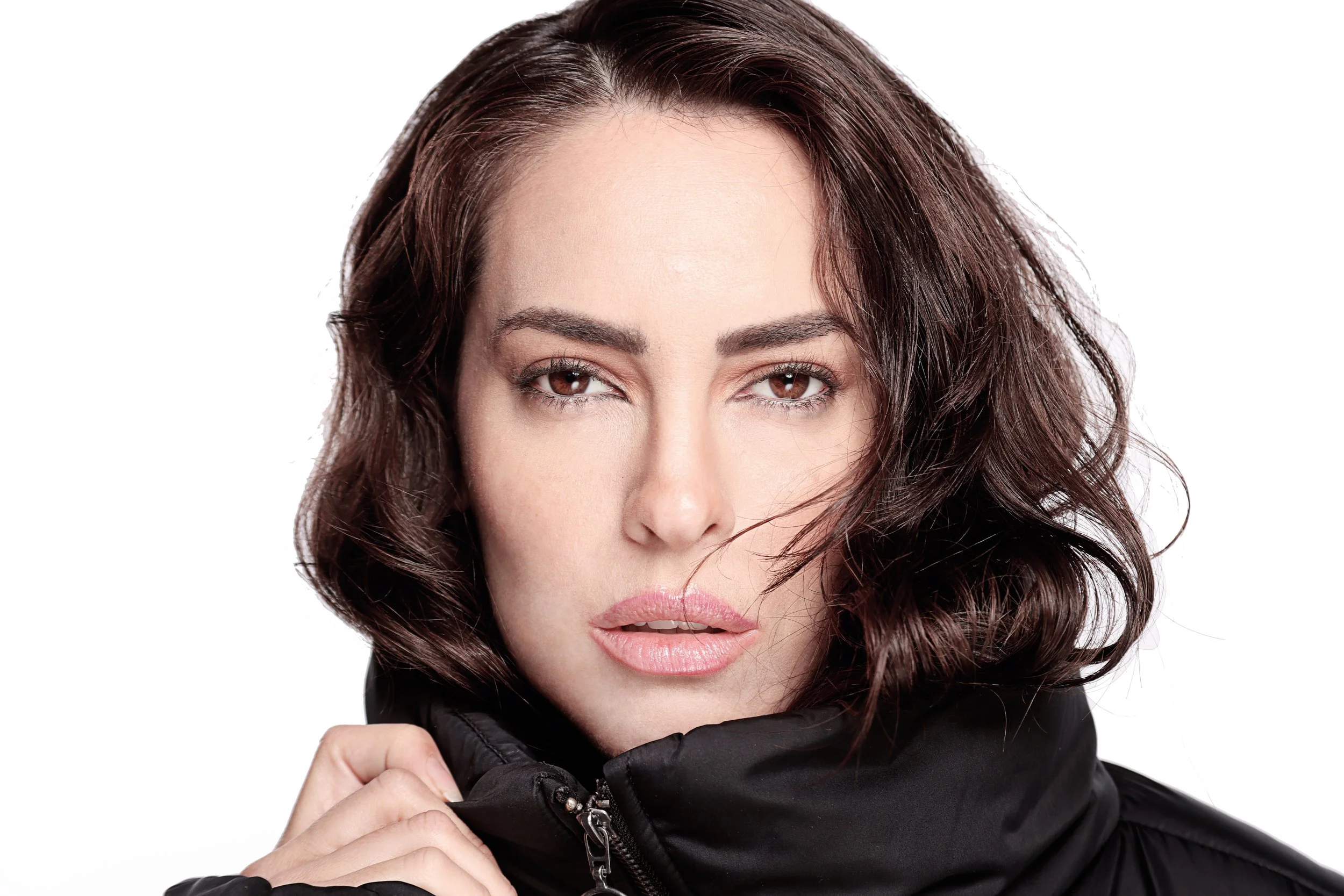 A woman with wavy brown hair and brown eyes wearing a black leather jacket, holding its collar with her hand, against a plain white background.