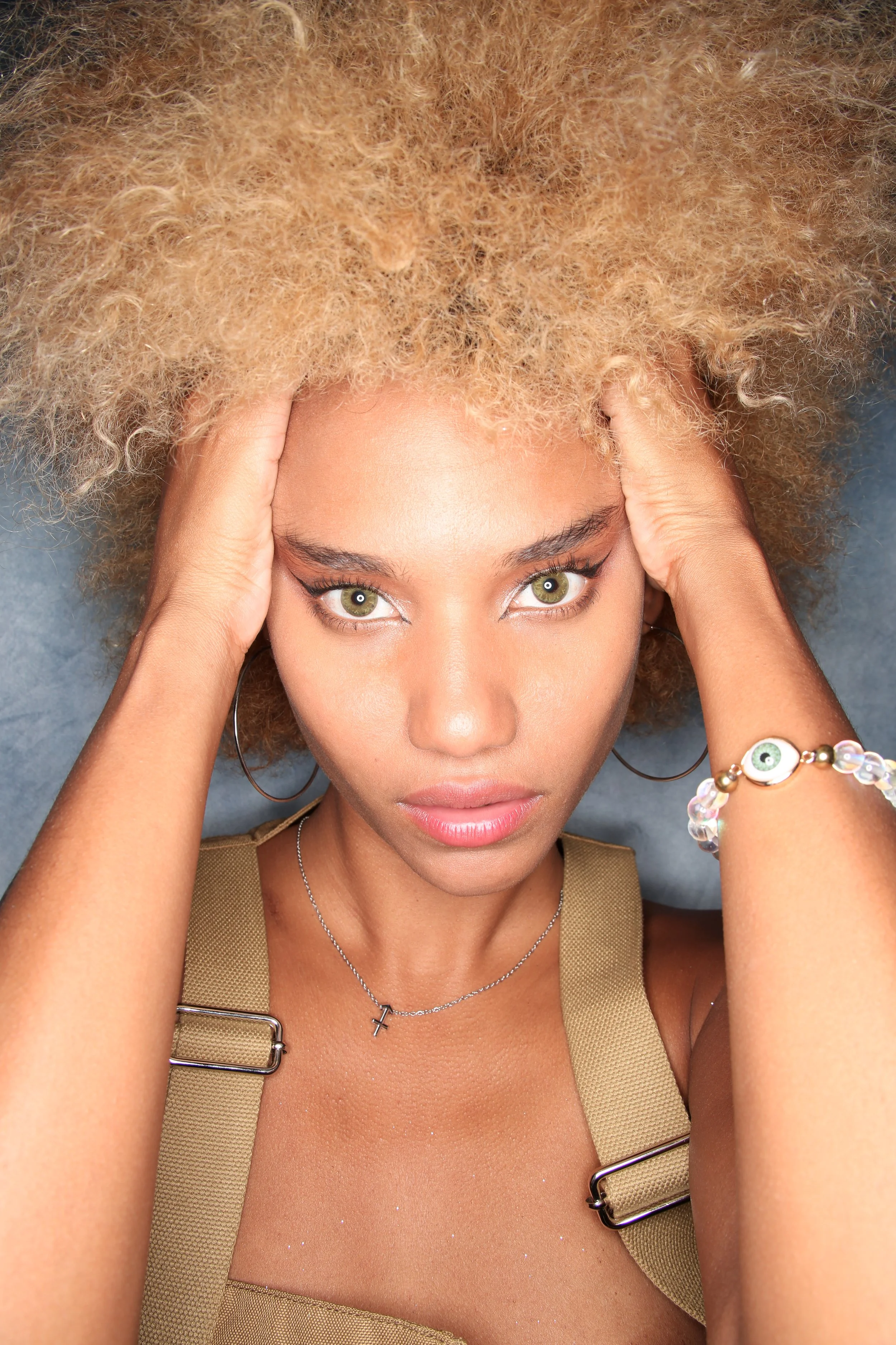 A woman with light skin and green eyes lying on her back, holding her head with both hands, hair spread out, wearing a beige tank top, jewelry including a cross necklace and a bracelet with eye charms.
