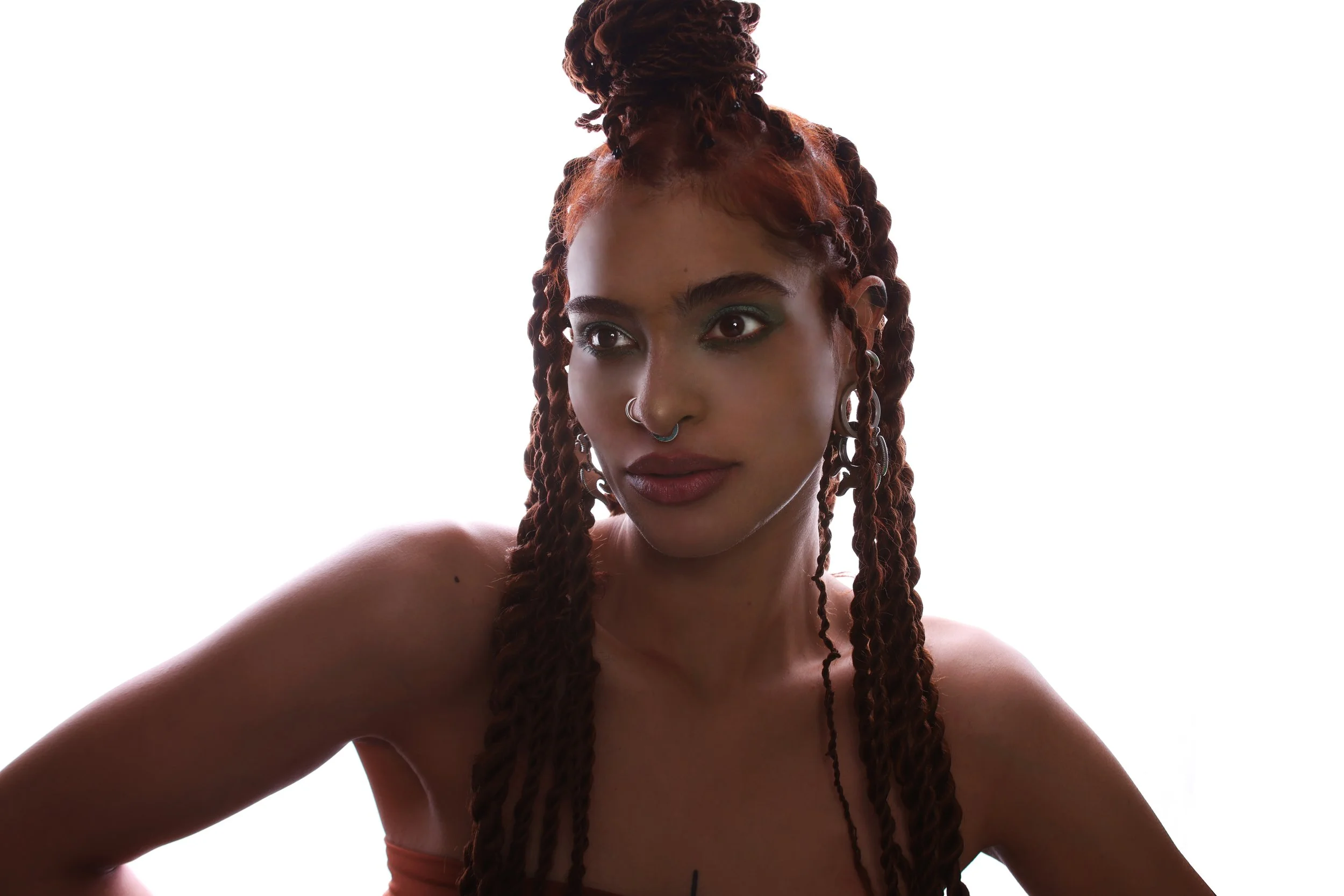 A woman with braided hair and multiple facial piercings, including nose and septum jewelry, looking off to her right against a white background.