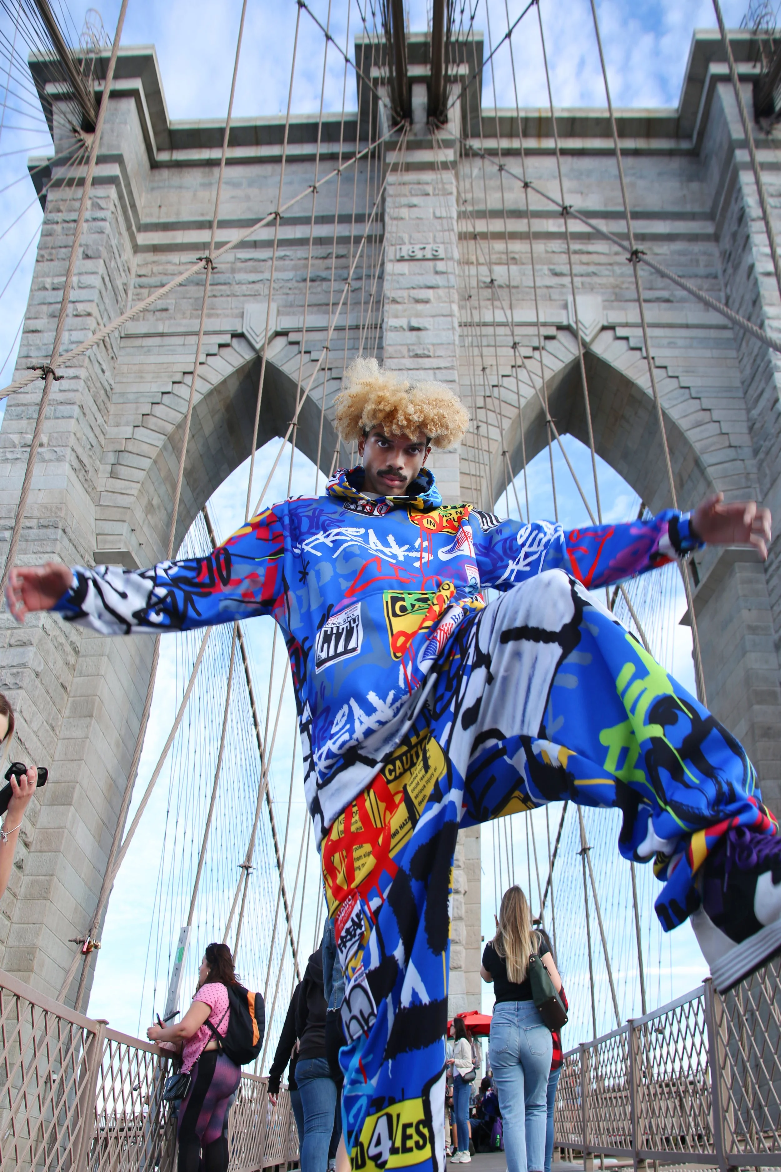 A young man with blonde curly hair and a mustache is standing on the Brooklyn Bridge in New York City, posing for a photo. He is wearing a colorful, graffiti-themed hoodie and pants. The bridge's stone towers and cables are visible in the background,