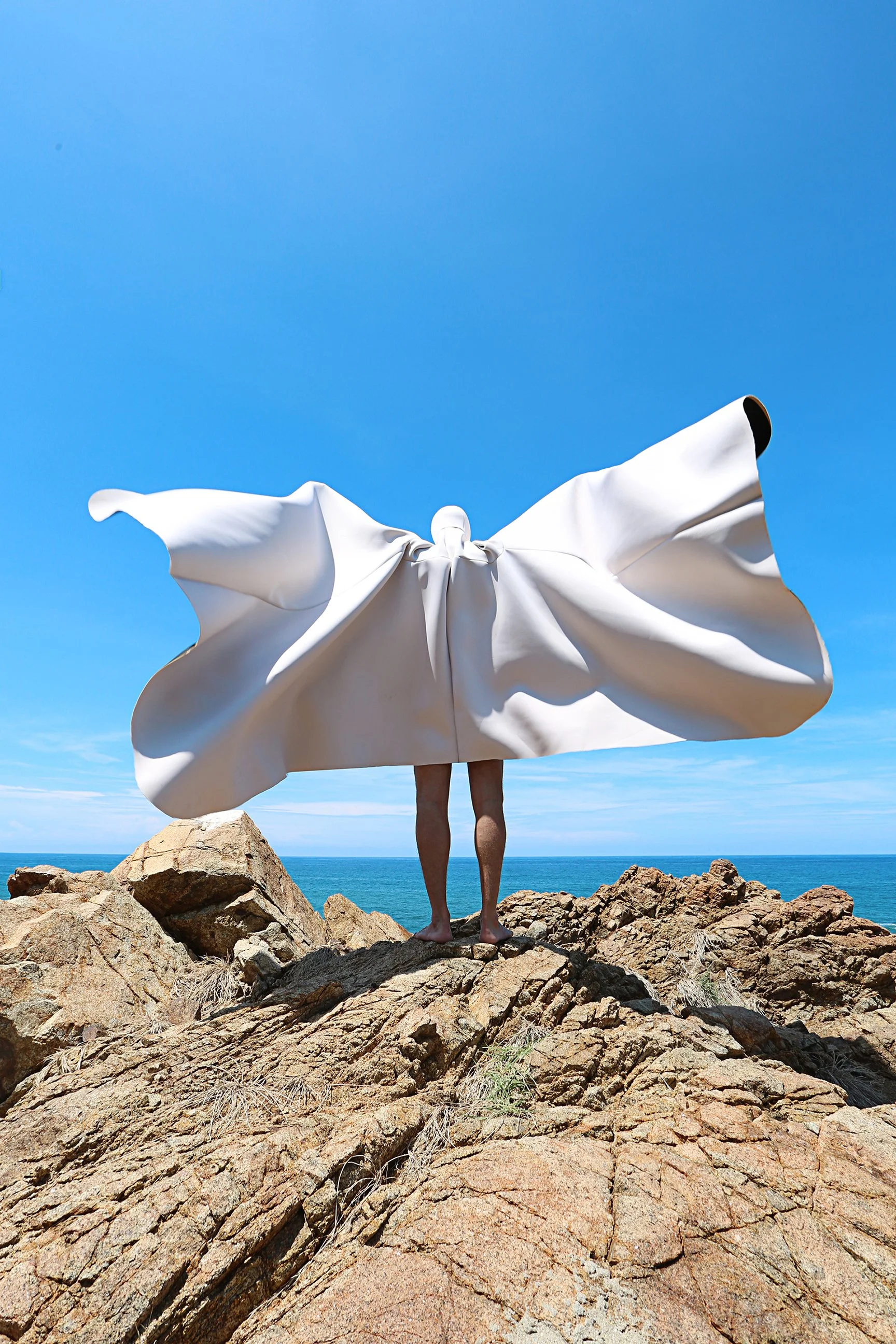 Person standing on rocky shoreline holding a large white flag or sheet, with the ocean and clear blue sky in the background.