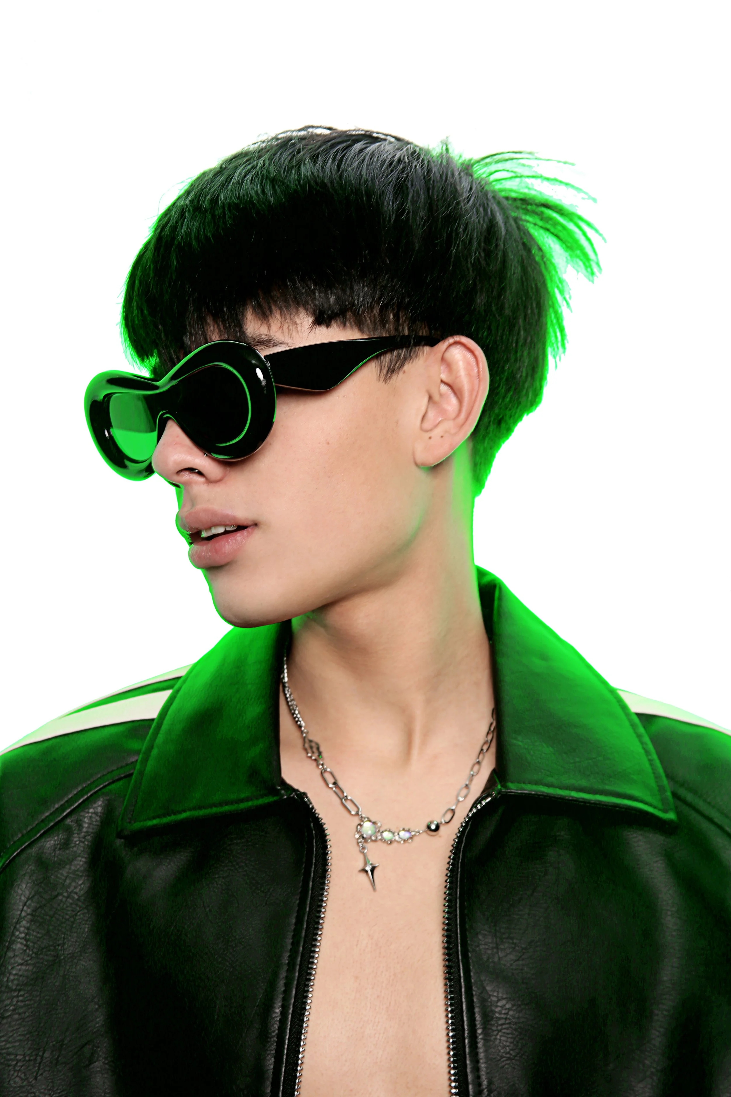 A person with short, black hair and green highlights wearing large, black sunglasses with green rims, a black leather jacket, layered necklaces, and standing against a white background.