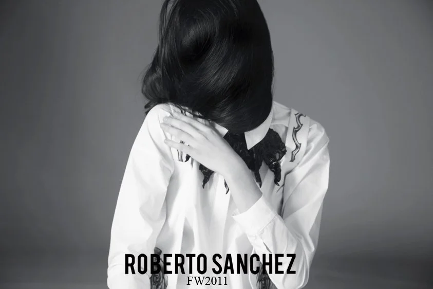 Black-and-white photo of a woman with dark hair covering her face, wearing a white blouse with black lace details, against a solid gray background. Text at the bottom reads 'ROBERTO SANCHEZ FW2011'.