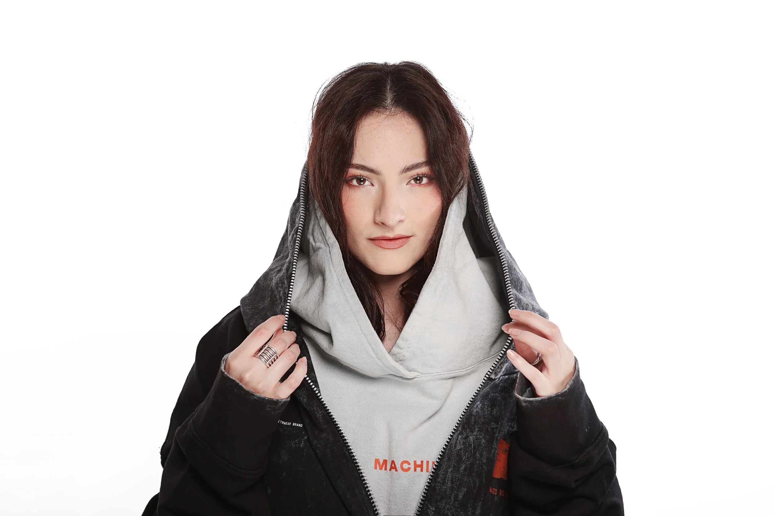 A young woman with dark brown hair and light makeup is lifting the zipper of her black hoodie, revealing a light gray inner hoodie with orange text. She is looking directly into the camera with a slight smile.