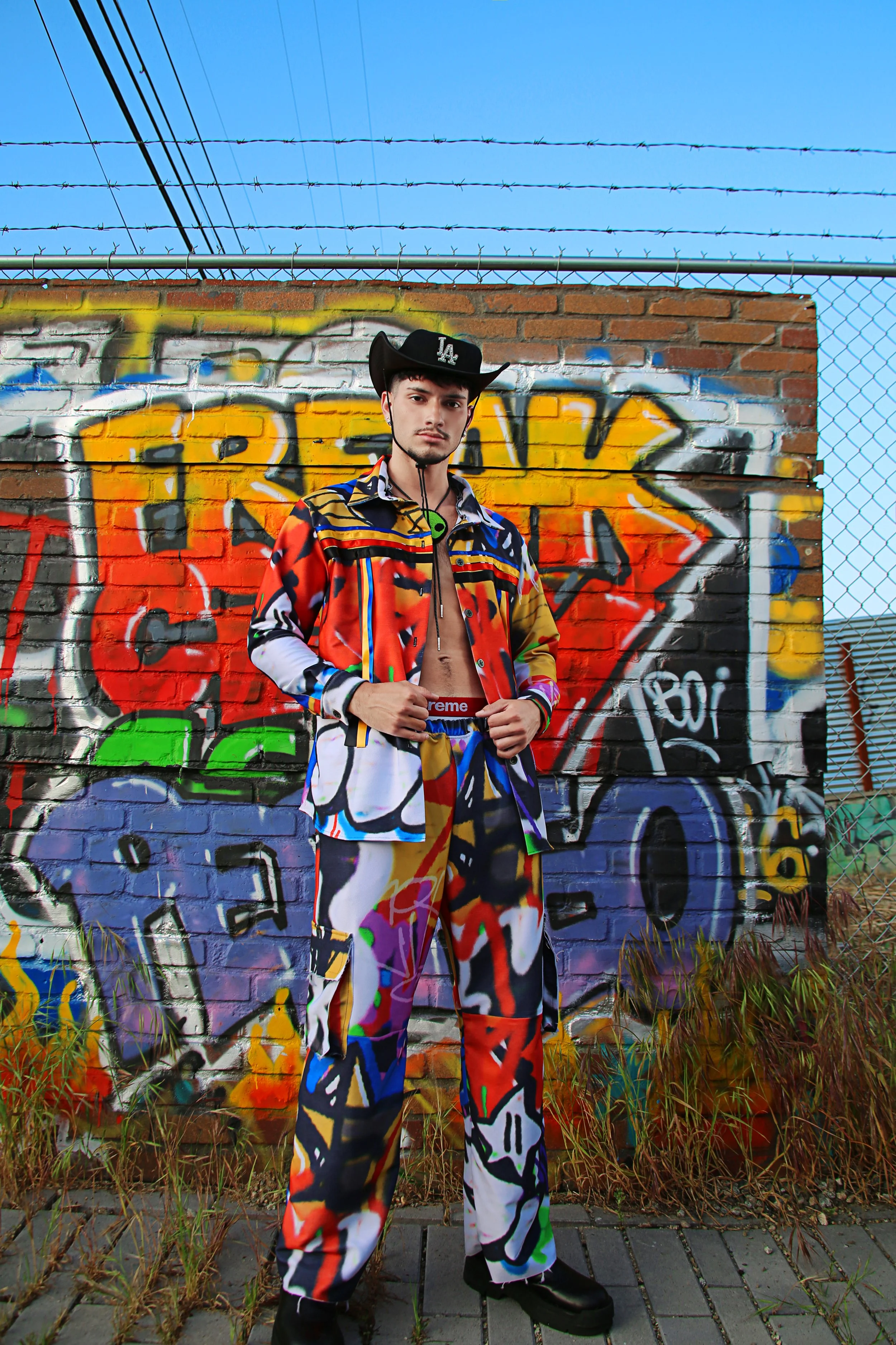 A young man standing in front of a graffiti-covered brick wall under a blue sky. He is wearing a multicolored, abstract-patterned jacket and pants, a black cowboy hat, and black shoes.