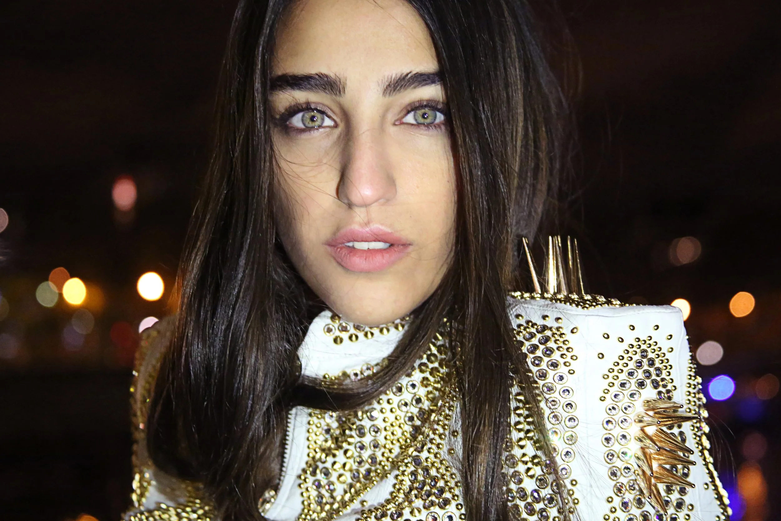 A young woman with long dark hair, green eyes, and makeup, wearing a white jacket with gold and clear jeweled embellishments, against a background of blurred colorful lights.