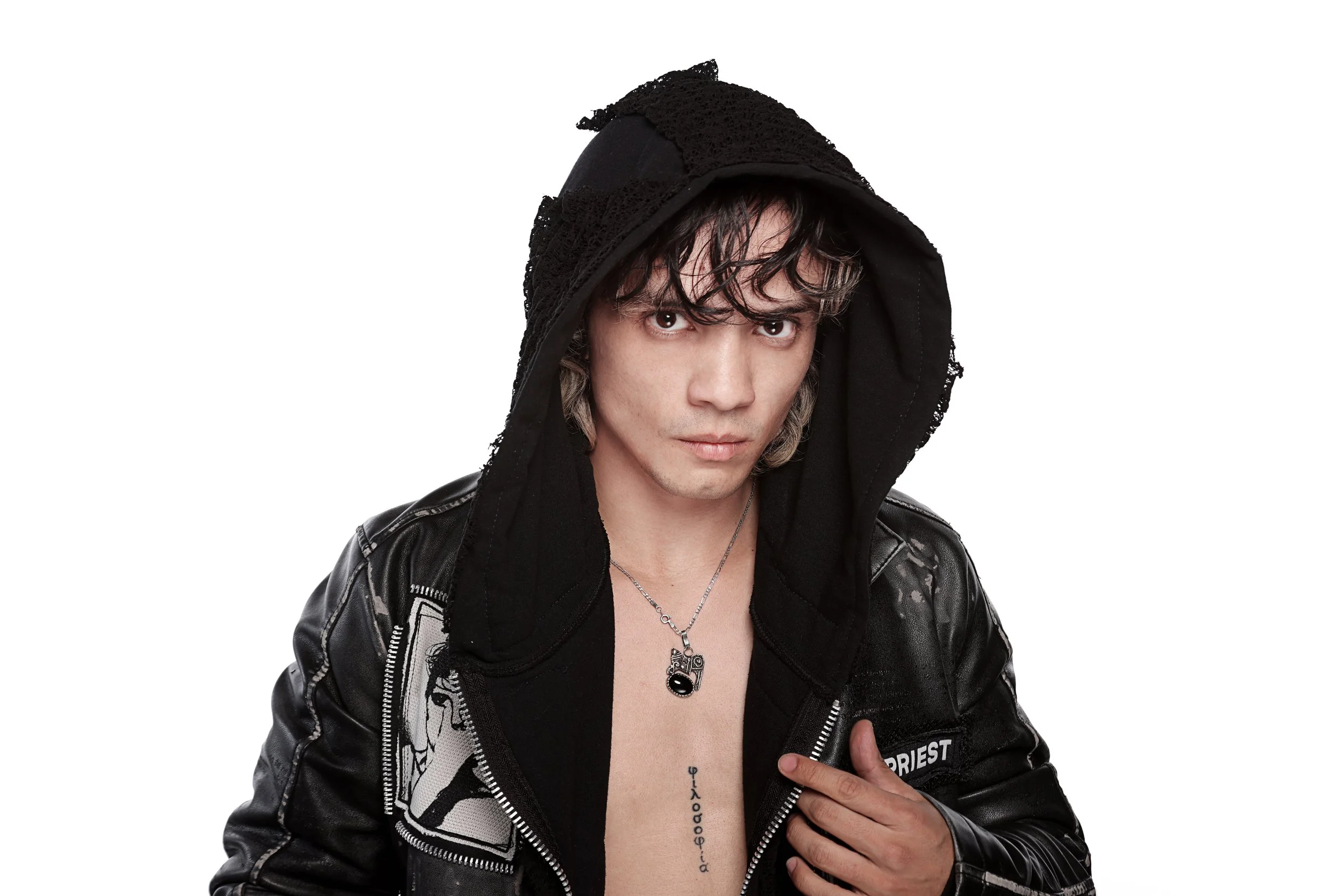 A young man with tousled dark hair, wearing a black hooded jacket with various patches, and a silver necklace with a pendant. He is looking directly at the camera with a serious expression, against a plain white background.