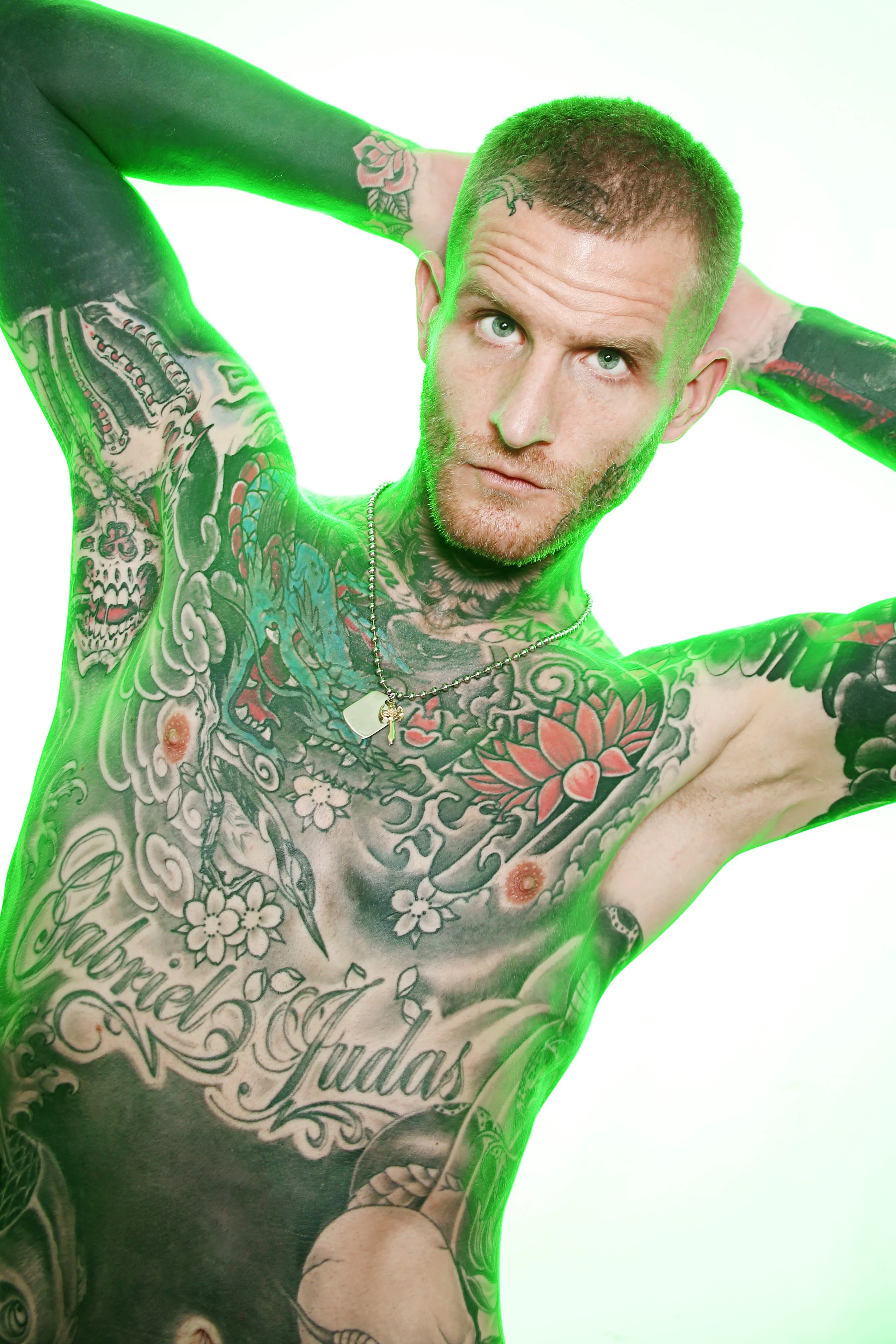 A tattooed man with short hair and light-colored eyes, shown from the chest up with his arms raised behind his head against a bright background with green light reflecting on his skin and tattoos.