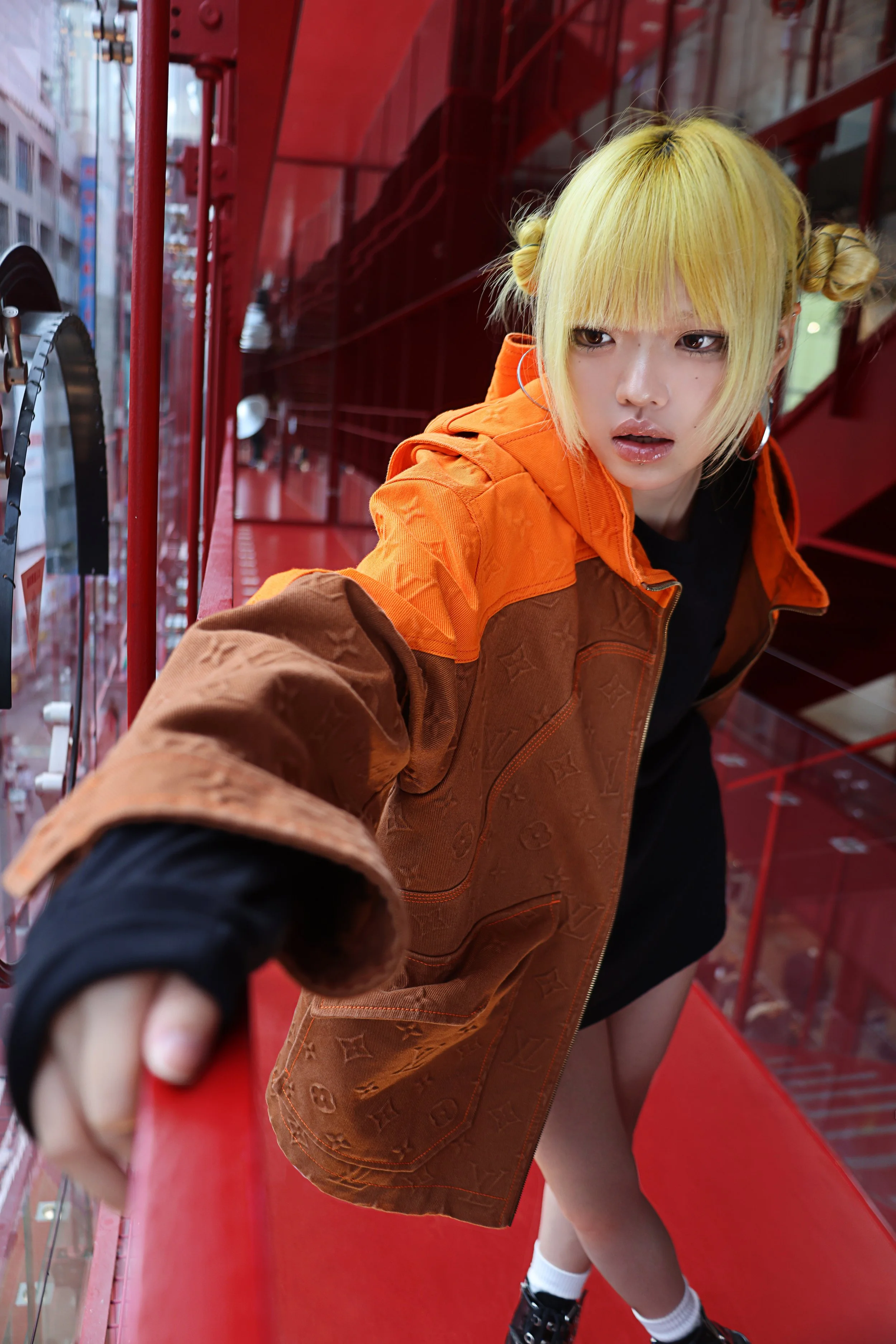 A person with blonde hair in pigtails leaning over a red railing on urban balcony, wearing an orange jacket, black shirt, white socks, and black shoes, with city buildings visible in the background.