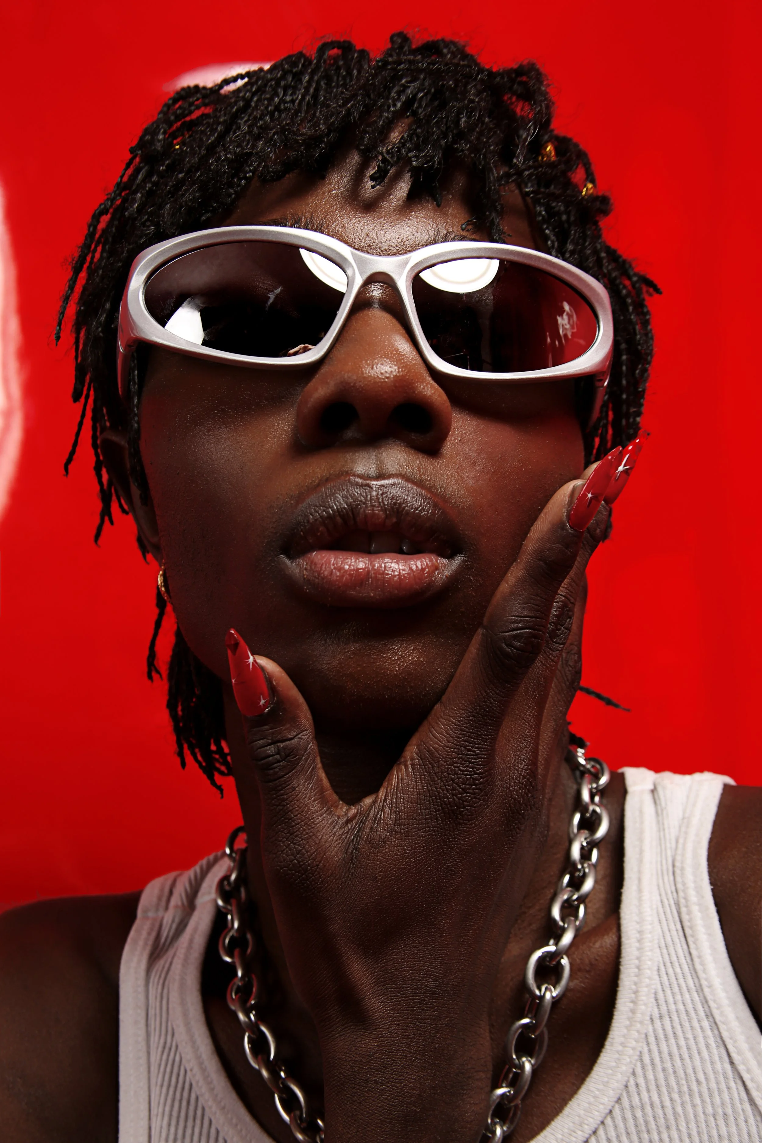 A person with dark skin wearing large white sunglasses, a silver chain necklace, and a white tank top, touching their face with their fingers against a red background.