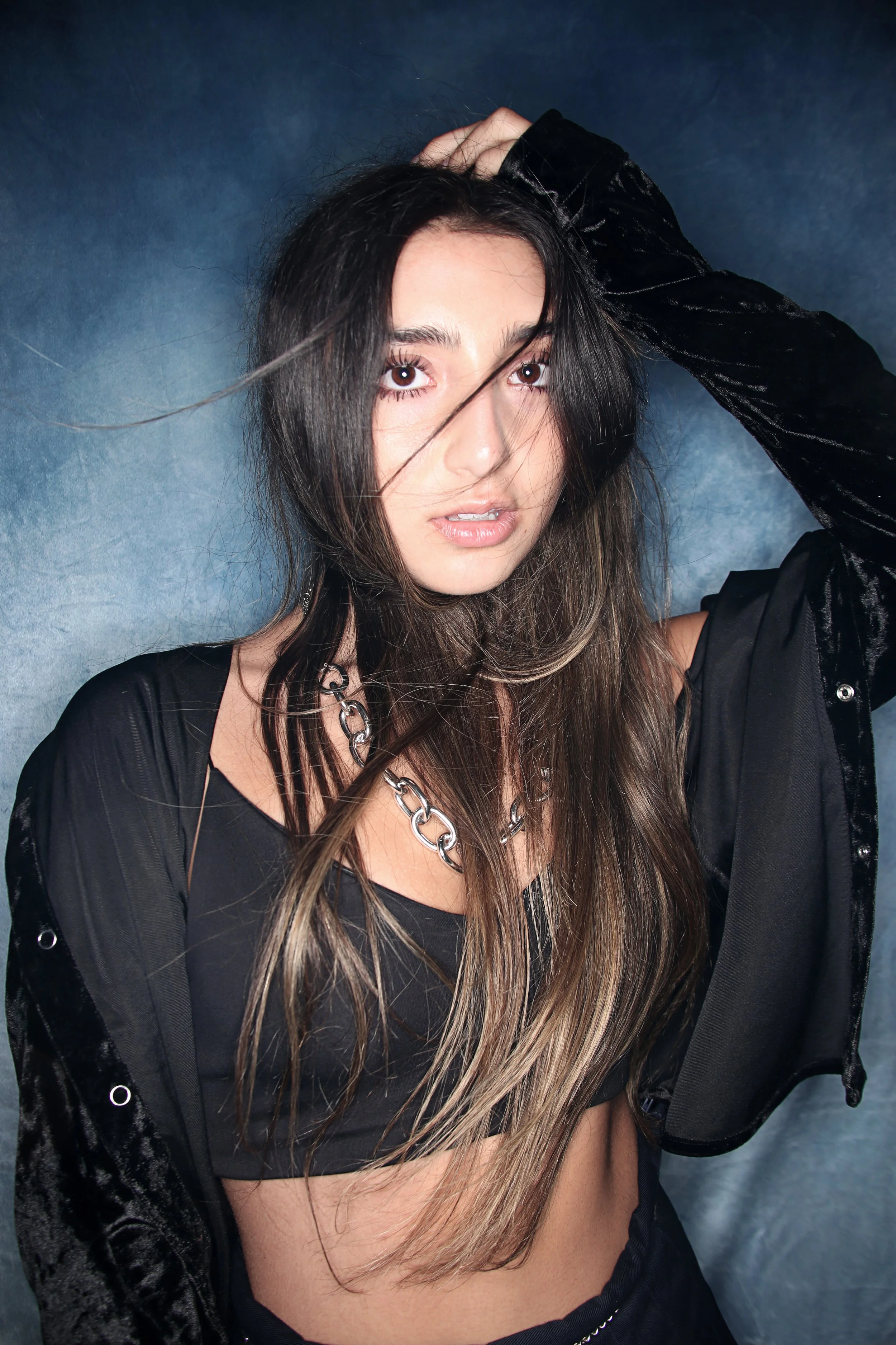 Young woman with long hair, wearing a black top and silver chain necklace, posing against a blue background, with one arm raised and hand on her head.