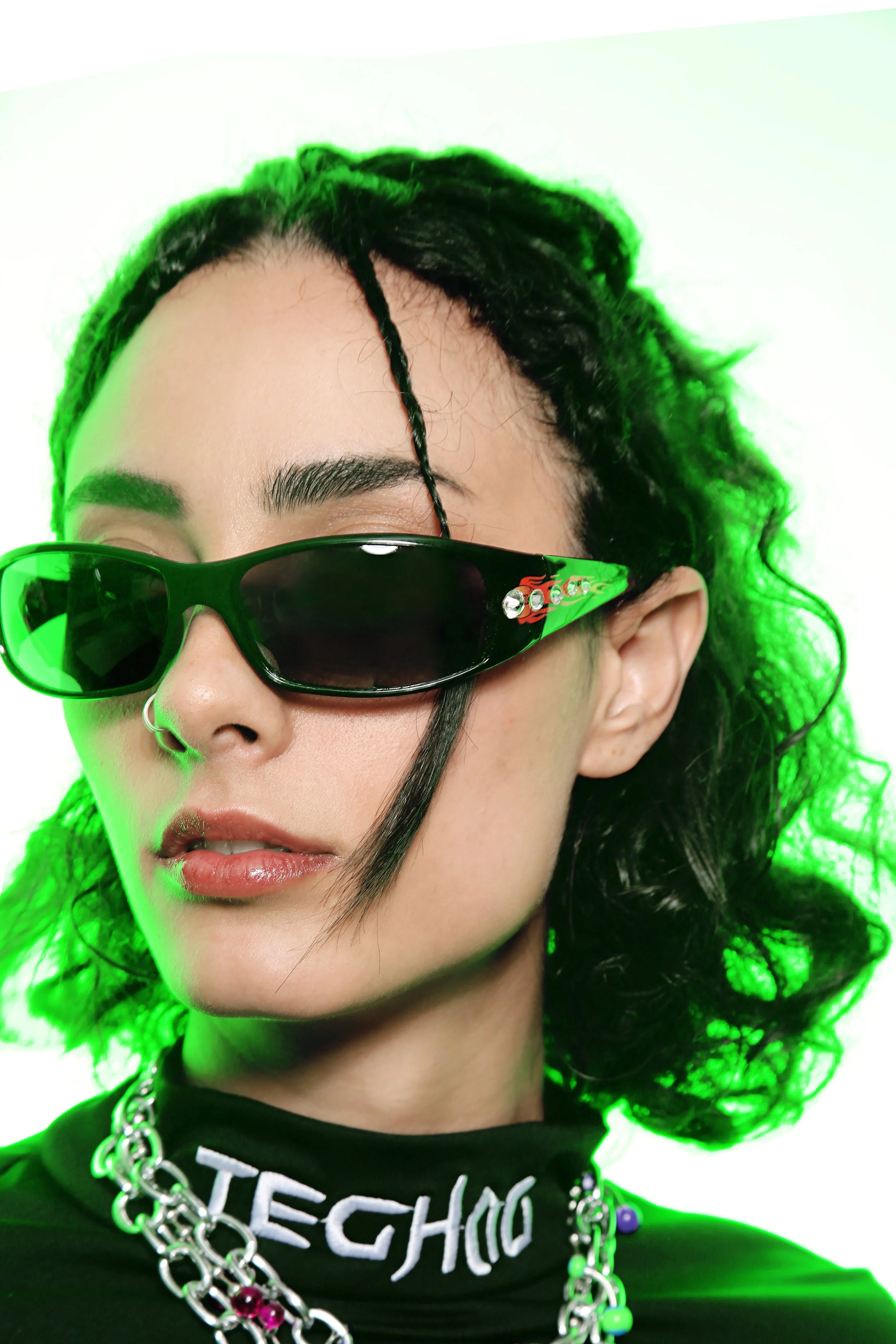 Close-up of a woman wearing black sunglasses with green lighting, black curly hair, and layered jewelry, including chains and a necklace with pink and blue stones. She is also wearing a black top with the word 'TECHNO' partially visible.