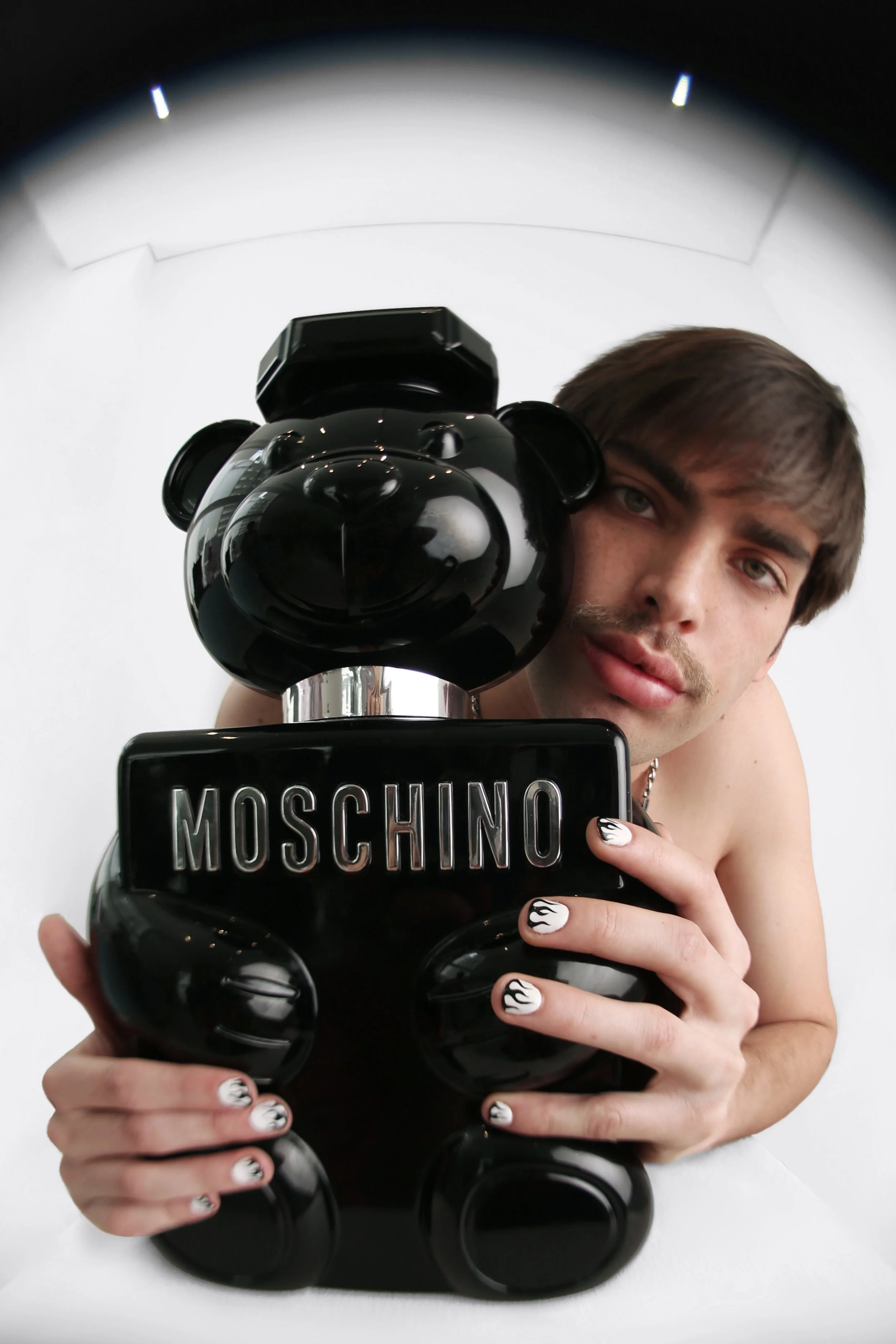 Young man taking a mirror selfie holding a black Moschino bear-shaped handbag in front of him, partially covering his face. The man has light skin, light brown hair, and painted nails with black and white flames.