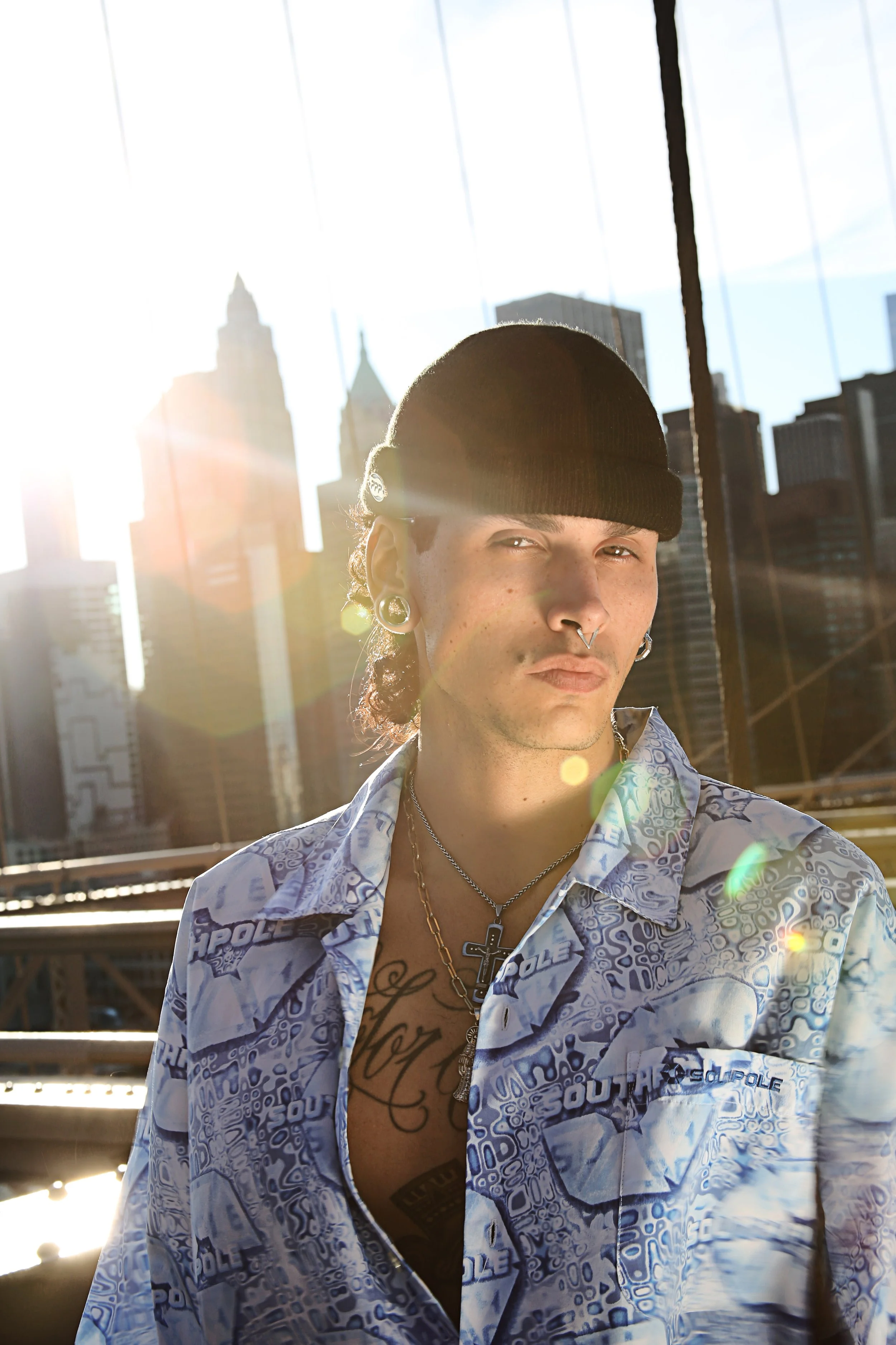 A young man wearing a black beanie, multiple earrings, and layered necklaces stands on a city rooftop with tall buildings in the background, backlit by the setting sun creating lens flare.