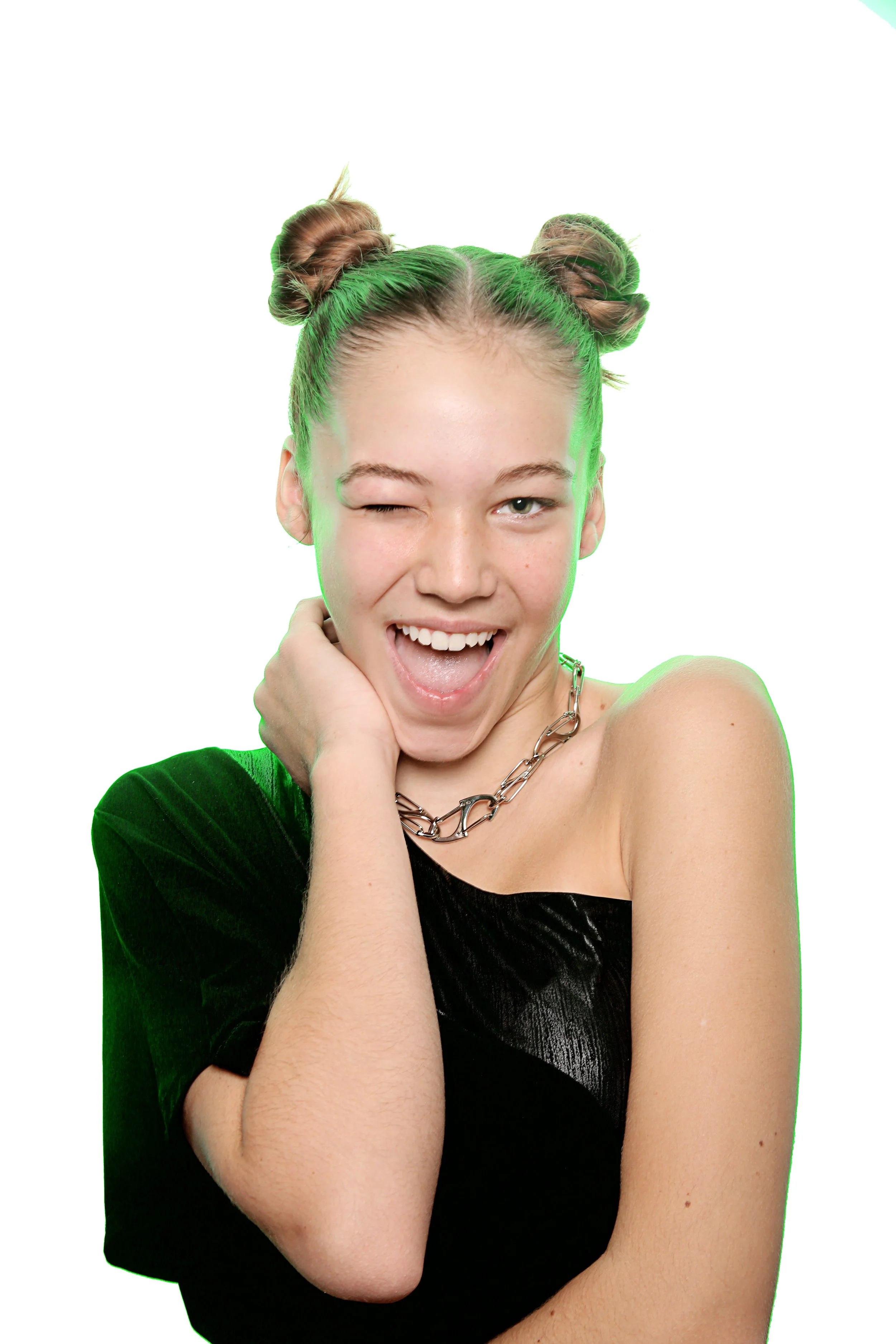 A young woman with braided hair styled into two space buns, wearing a black off-shoulder top, necklaces, and making a playful expression with one eye closed and mouth open, against a white background.