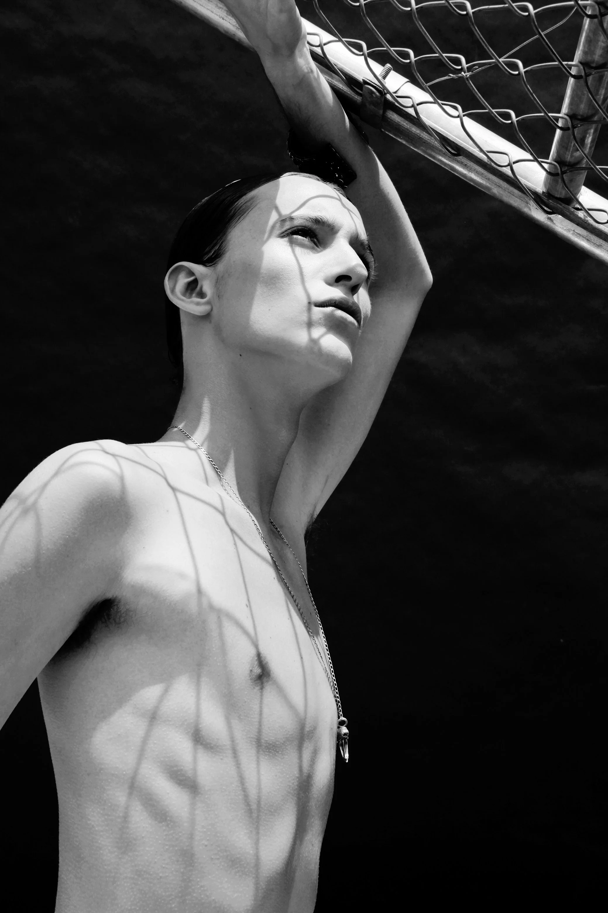 Black and white photo of a shirtless woman with wet hair, raising her arm over her head, with shadows cast on her body from a chain-link fence.